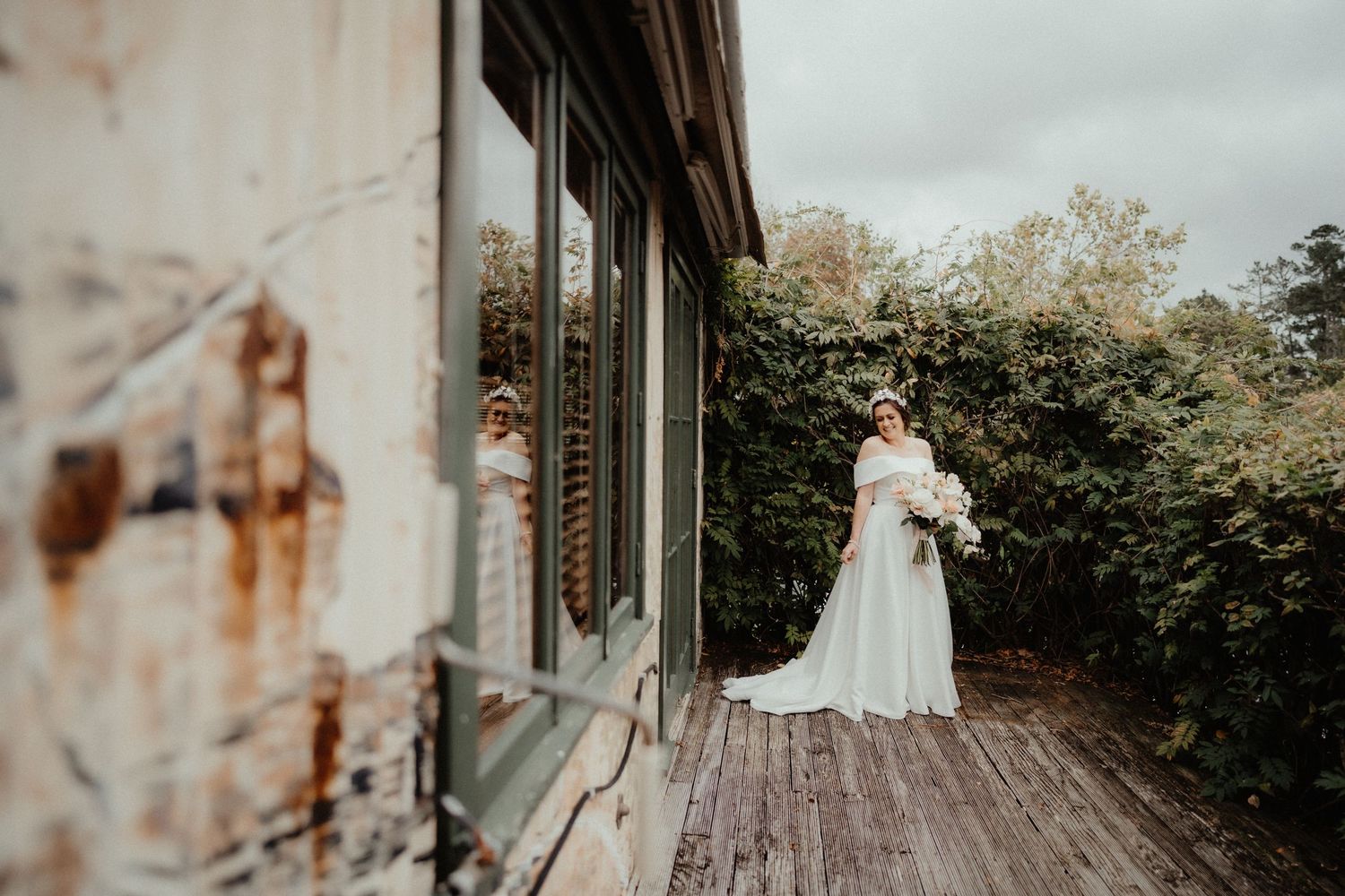 Bride in a long white gown holding a bouquet stands on a rustic wooden deck beside a stone building and green hedge.