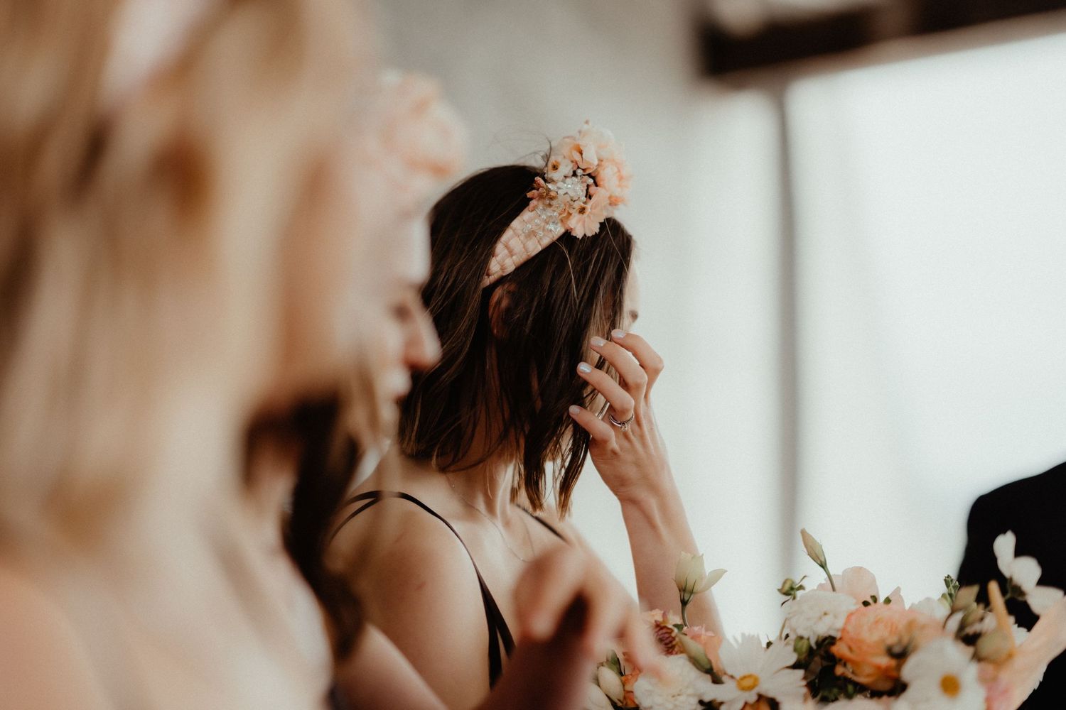 Bridesmaid with a floral hair piece wipes her eye during emotional wedding speeches beside a flower arrangement.