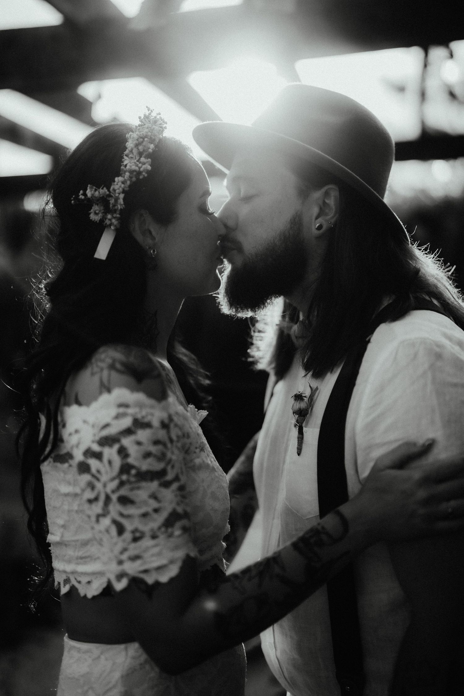 Black and white photo of a bohemian bride with a flower crown kissing her bearded, tattooed groom in a hat.