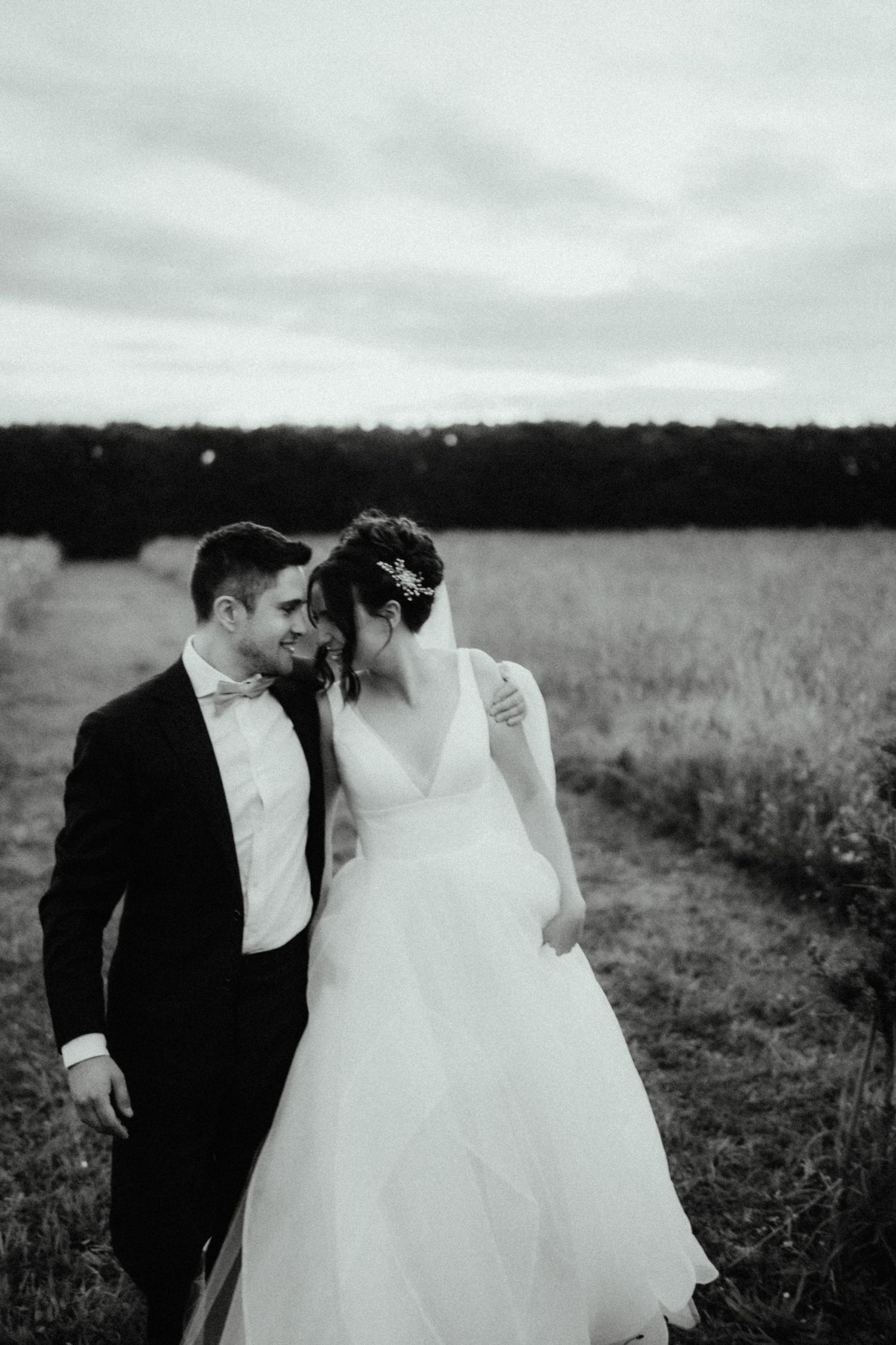 Black and white image of a smiling groom in a suit nuzzling his bride in a tulle gown as they walk through a grassy field.