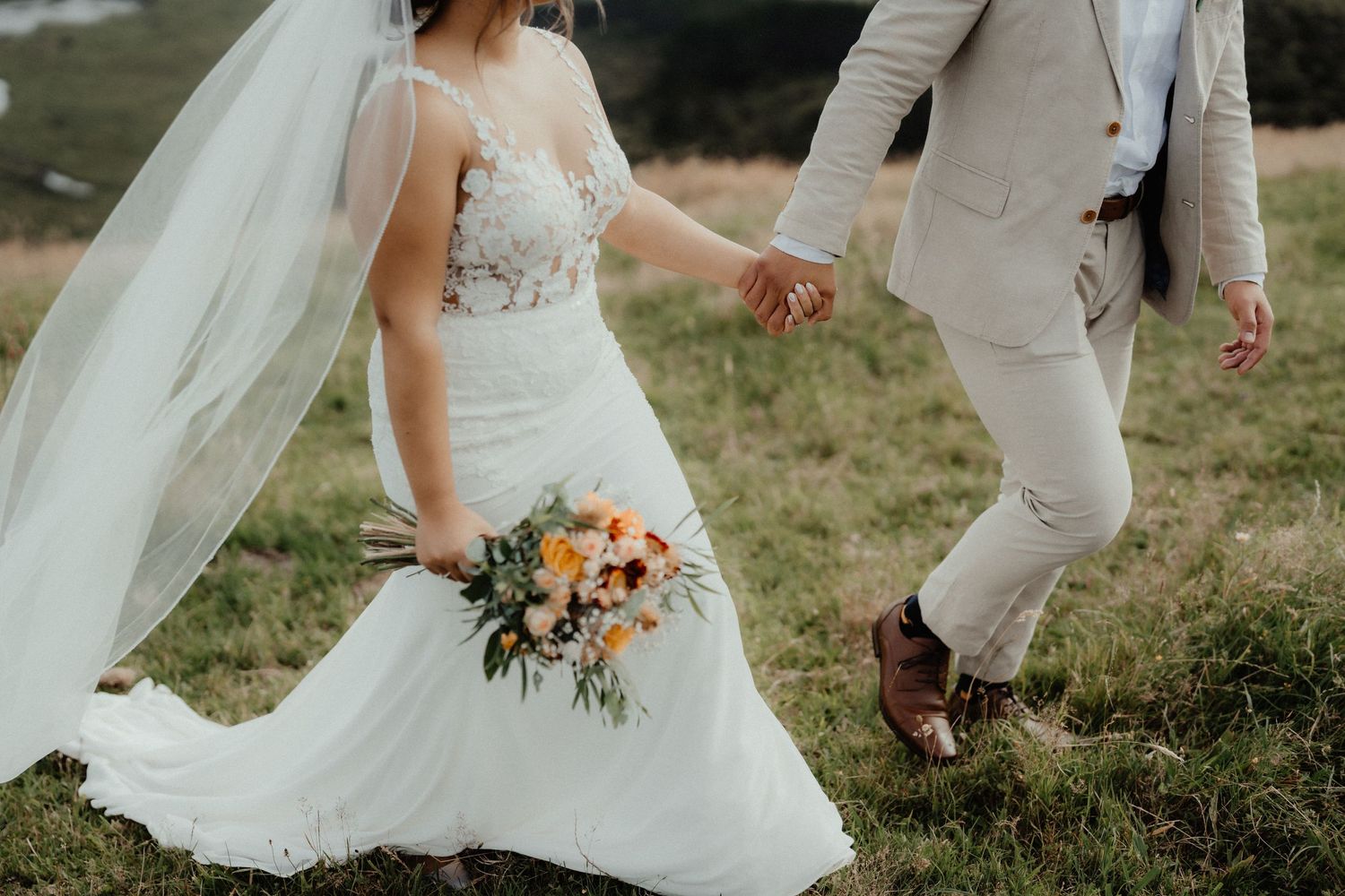 Bride in a lace wedding gown and long veil holds a colorful bouquet while walking hand in hand with the groom across a grassy field.
