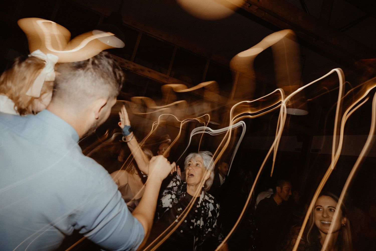 Wedding guests dance and celebrate on the dance floor, with motion-blurred light trails and an older woman smiling at the camera.