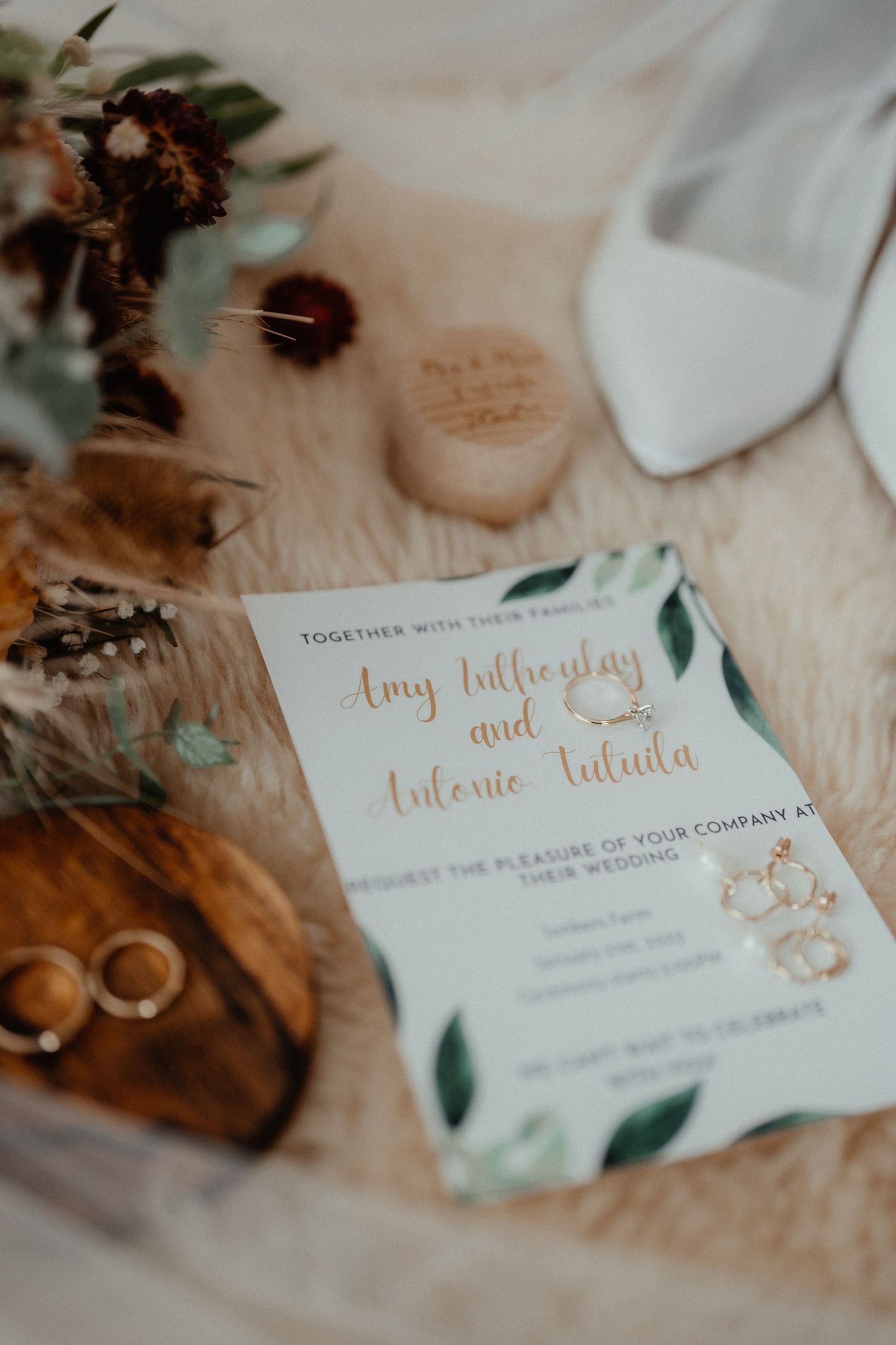 A wedding invitation for Amy and Antonio rests on a fur rug beside bridal shoes, gold rings, dried flowers and a wooden ring dish.