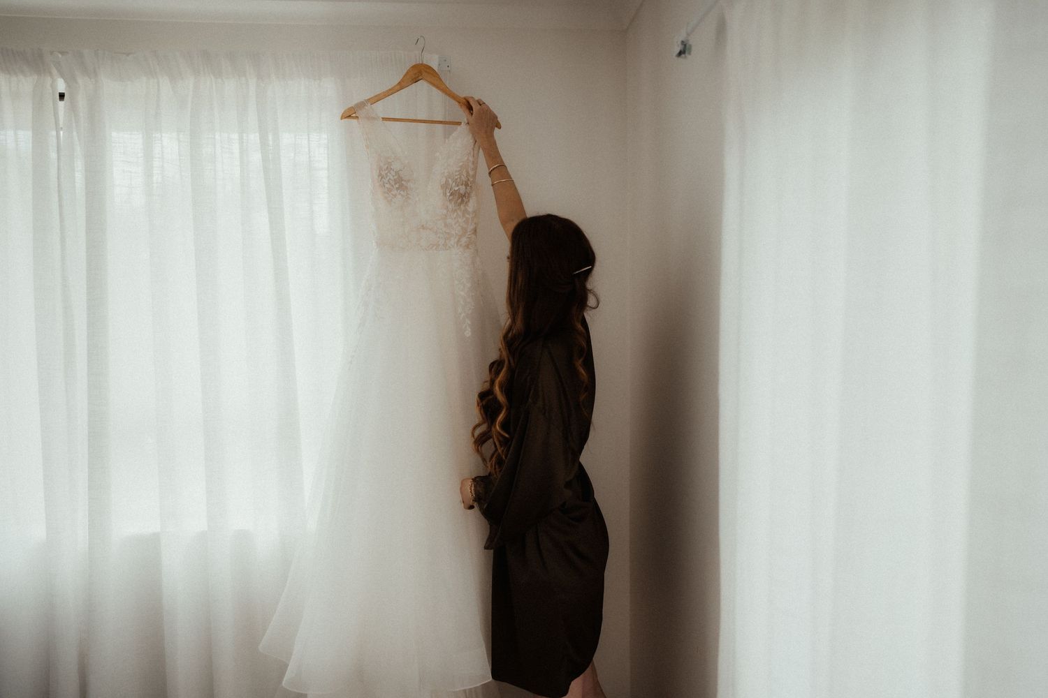A woman in a satin robe reaches up to a white wedding dress hanging by a sheer-curtained window as she prepares for the wedding.