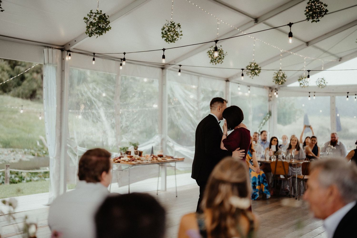 A couple dances at a wedding reception inside a marquee with hanging greenery and string lights while guests look on.