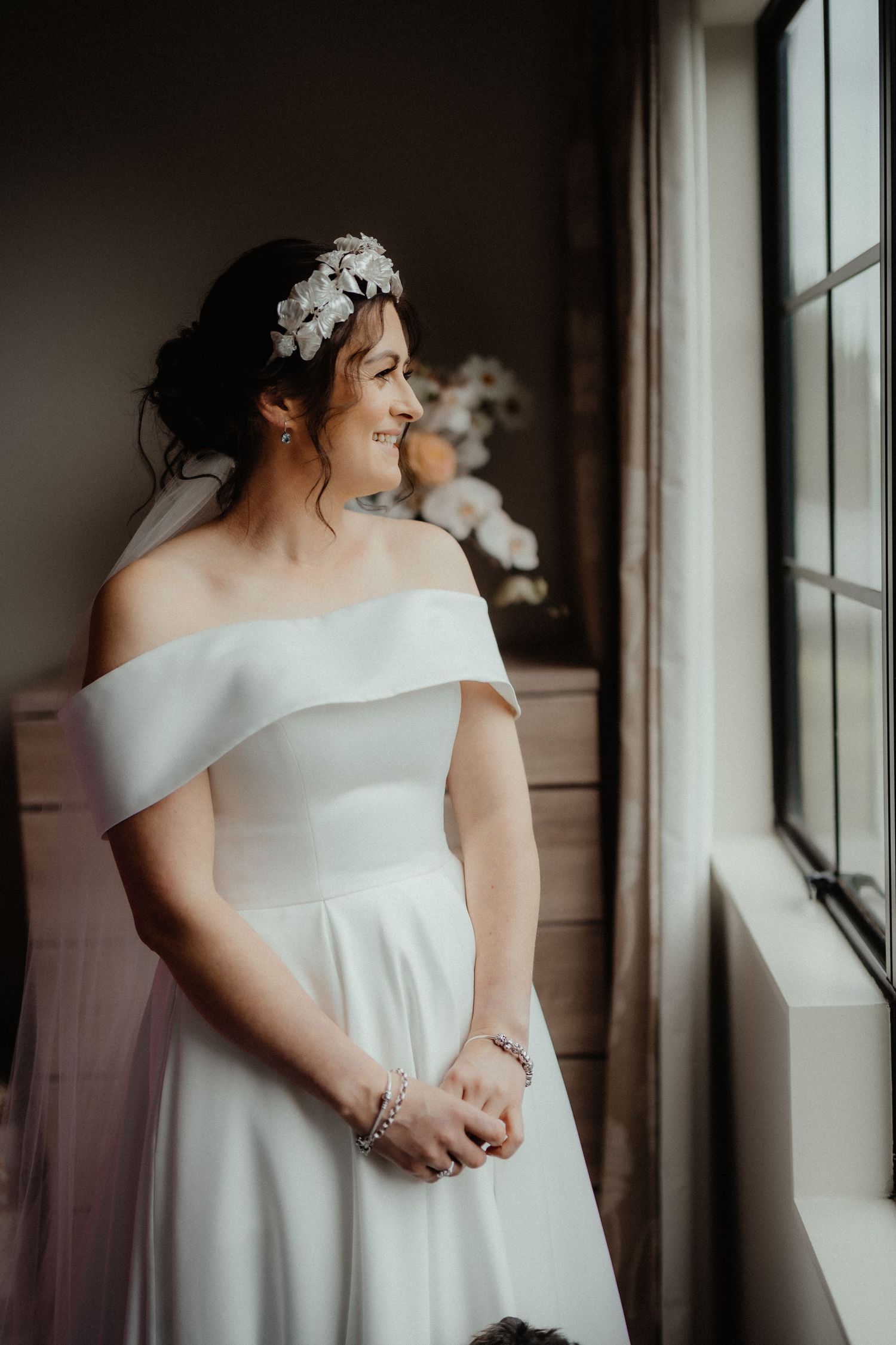 Smiling bride in an off-shoulder white gown and floral headpiece gazes out a window beside a soft floral arrangement.