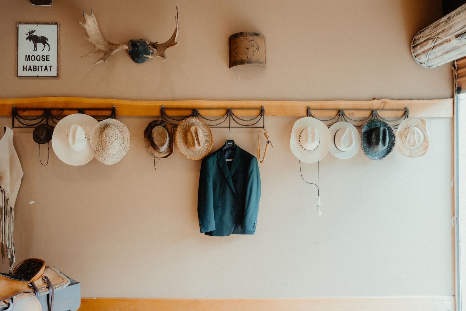 Rustic wall with a moose habitat sign, antler mount, cowboy hats on a wooden rack and a green suit jacket hanging beside them.