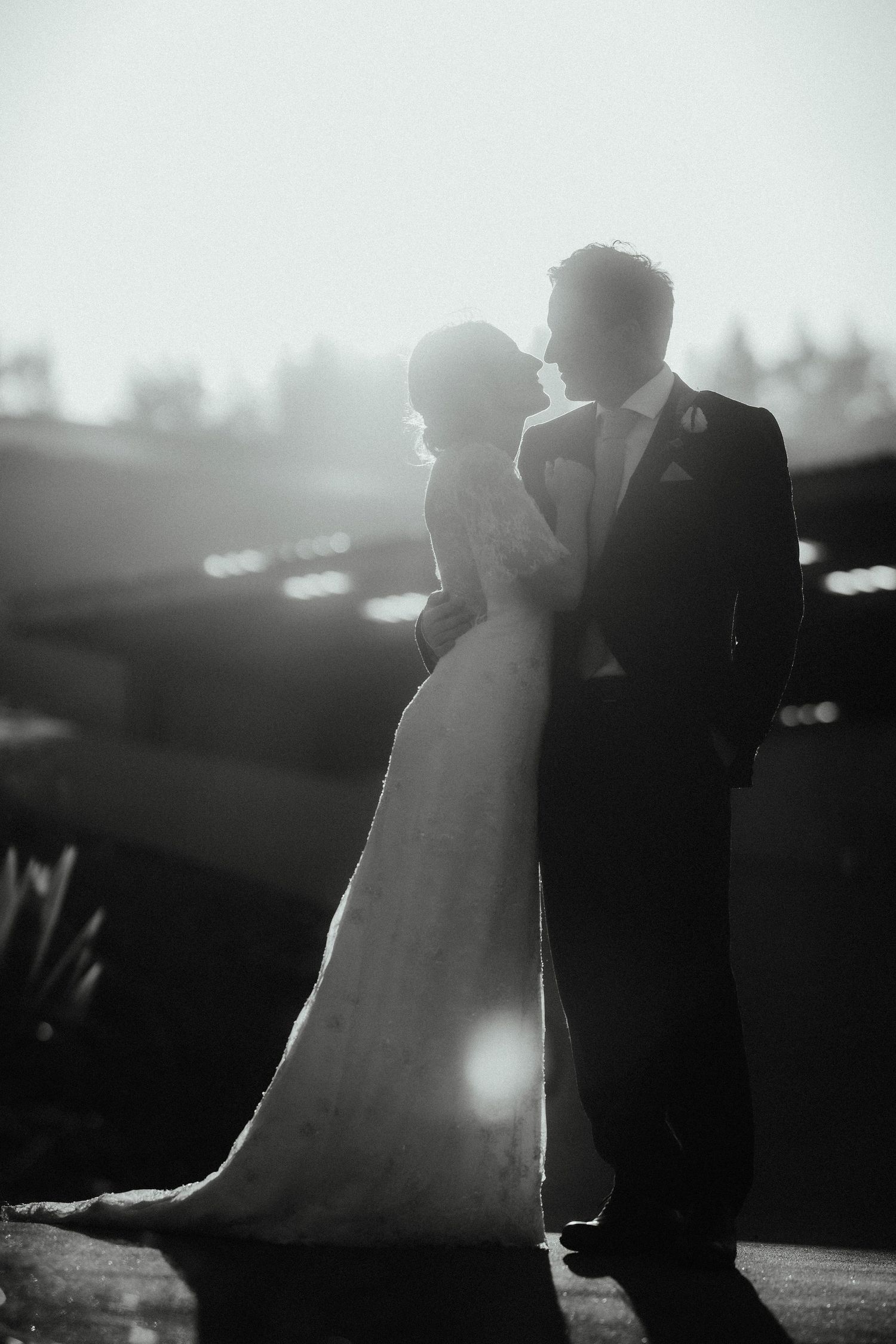 Black and white backlit silhouette of a bride and groom embracing and about to kiss at a countryside wedding venue.