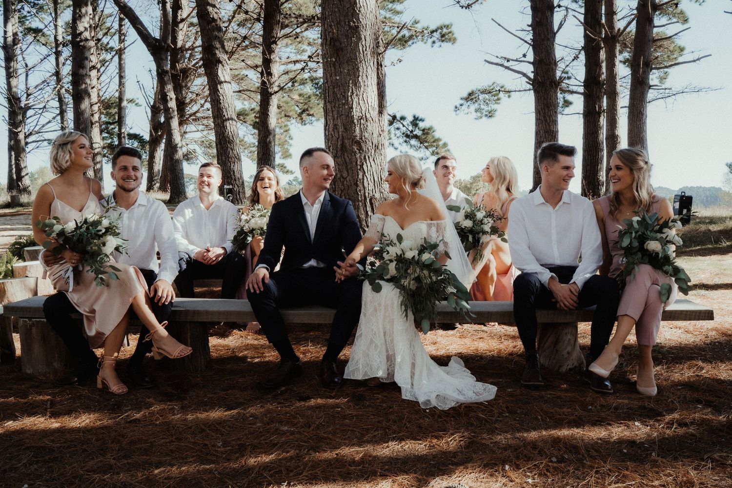 Bride and groom sit on a bench with bridesmaids and groomsmen, laughing together on pine needles in a sunlit forest.