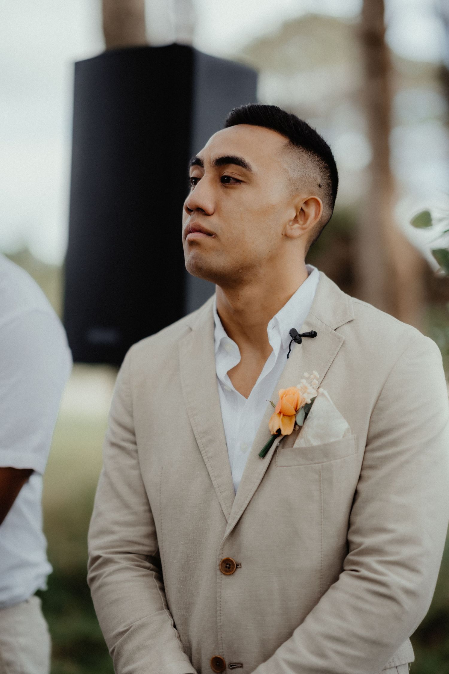 Groom in a beige linen suit with an orange boutonniere looks on solemnly during an outdoor wedding ceremony among the trees.