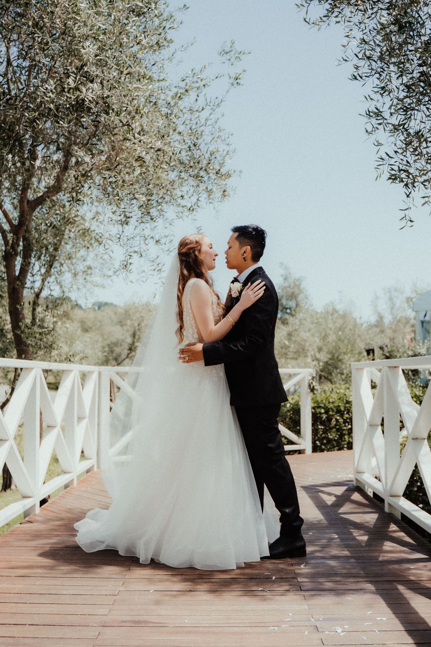 Bride in a flowing tulle gown and groom in a black suit embrace on a wooden deck with white railings and olive trees behind.
