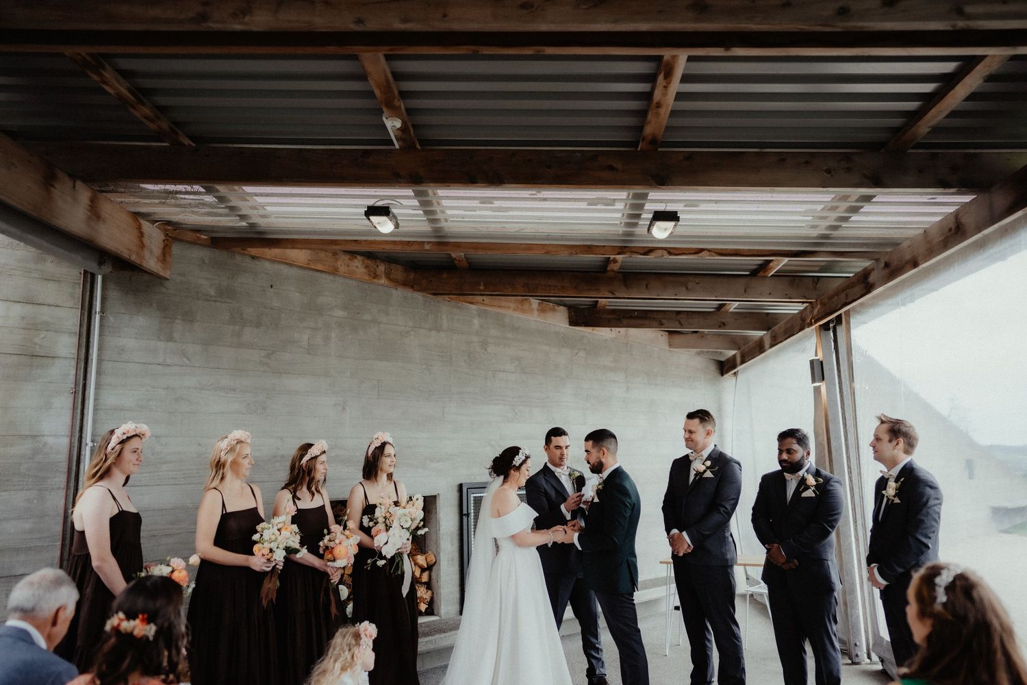 Bride and groom hold hands facing each other during an outdoor ceremony, with bridesmaids and groomsmen standing beside them.