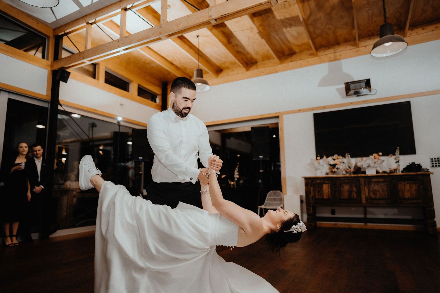 Groom dips the bride in a dramatic dance move beneath a timber-beamed ceiling at their wedding reception.