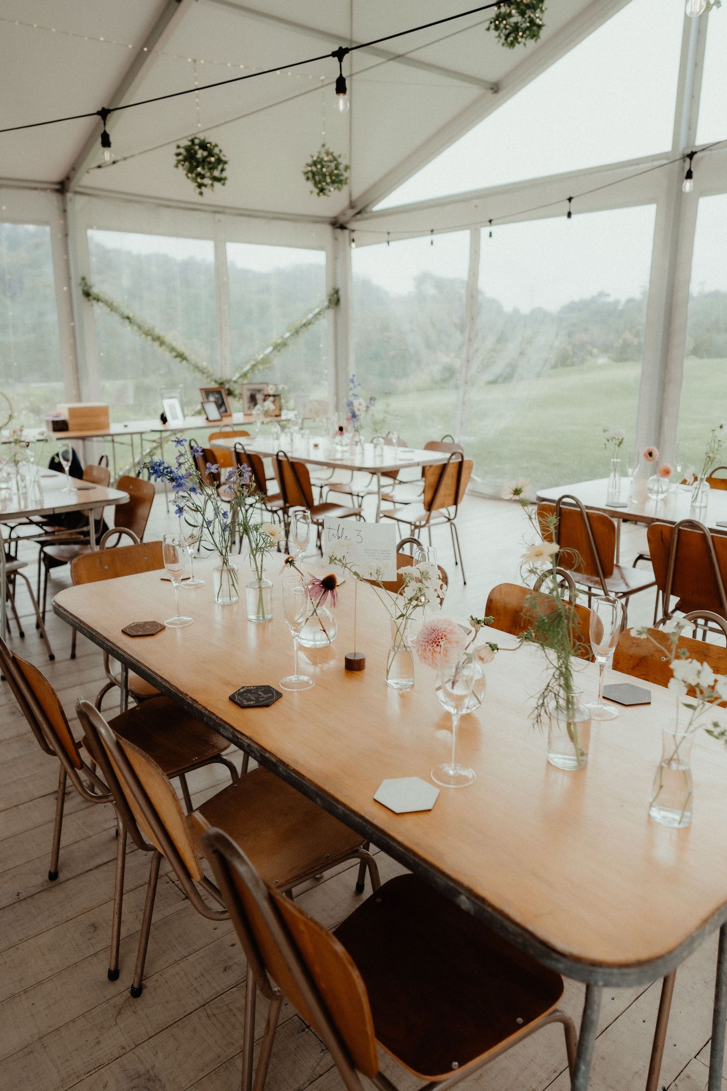 Wedding marquee with long wooden tables, leather chairs, string lights and small floral arrangements overlooking green hills.