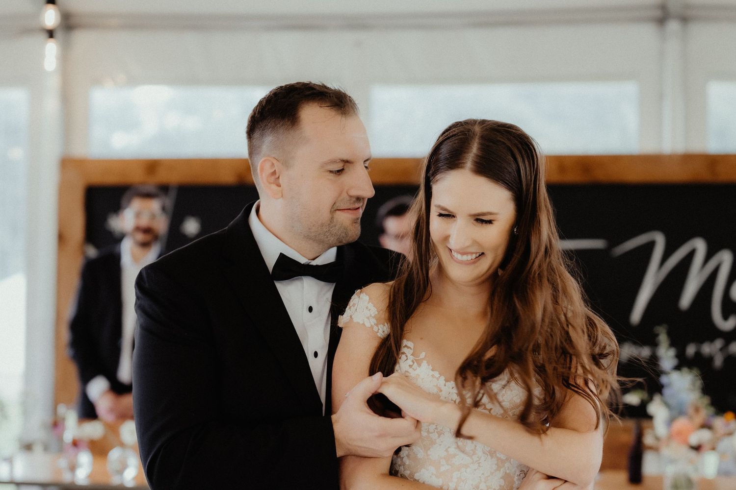Groom hugs his smiling bride from behind inside a reception tent as she looks down with joy.