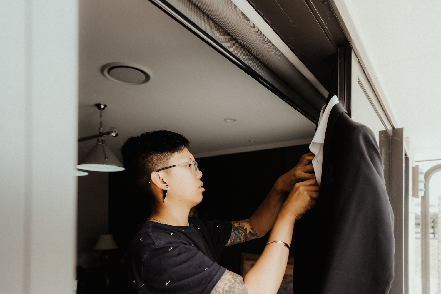 Man in glasses carefully adjusts a tuxedo jacket hanging near a window while preparing for a wedding.