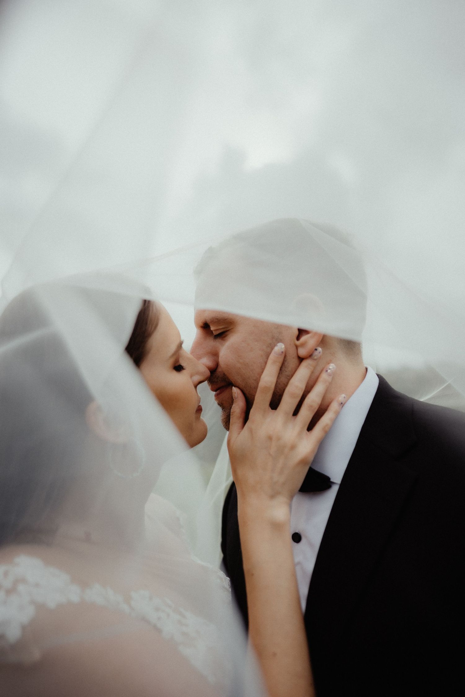 Bride and groom share an intimate kiss beneath a sheer white veil, her hand gently resting on his cheek.