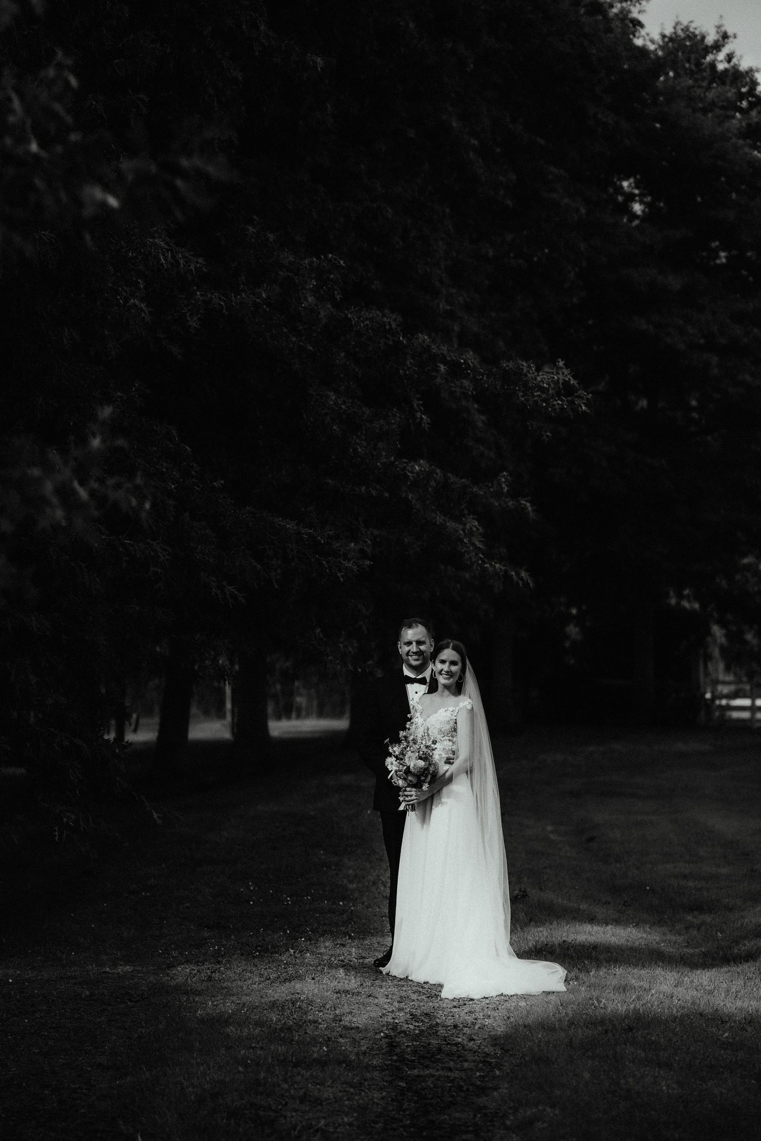 Black and white portrait of a smiling bride holding a bouquet and groom embracing her beneath tall dark trees.