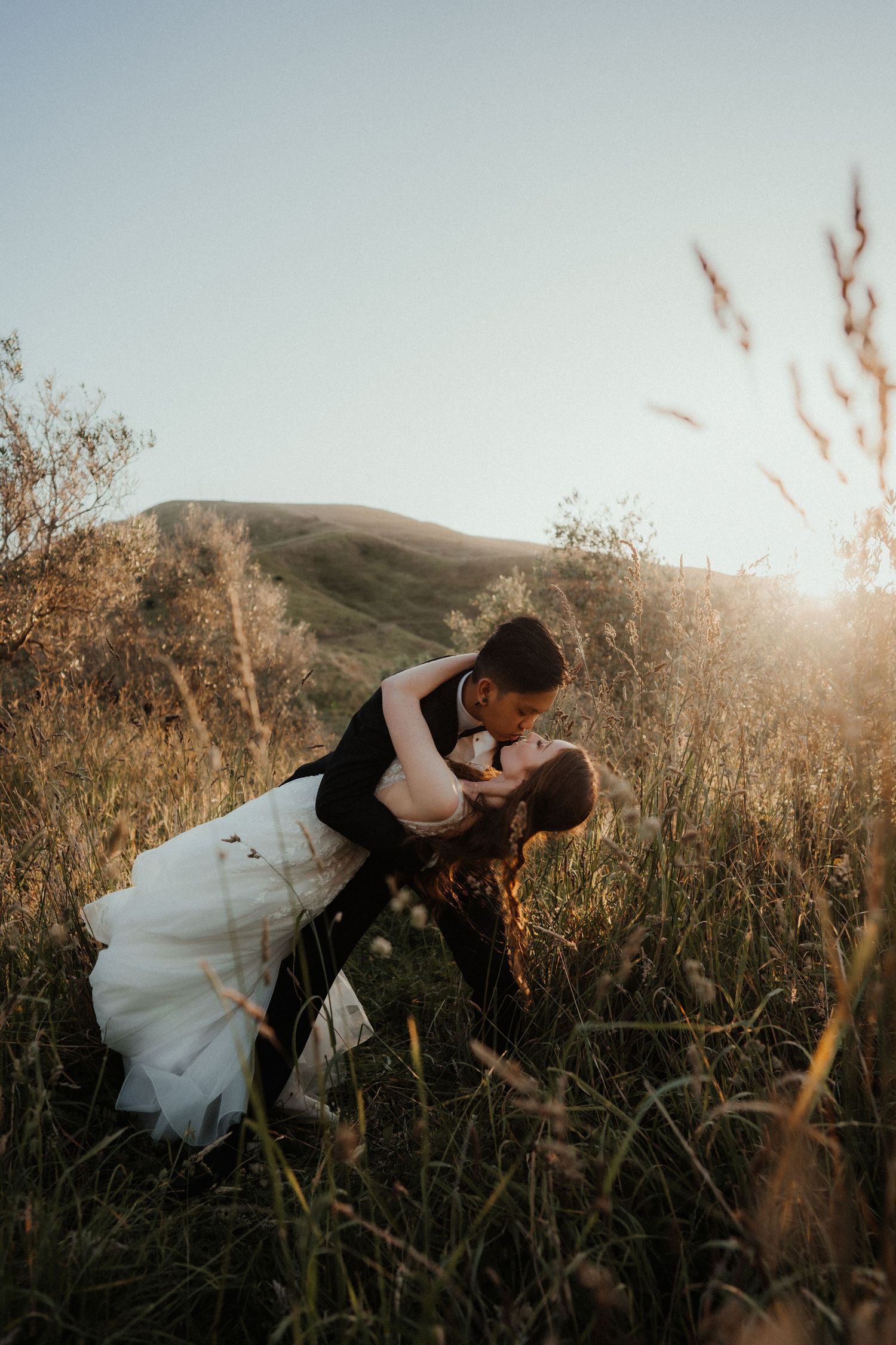 Groom dips and kisses his bride in a field of tall grass at sunset with rolling hills in the background.