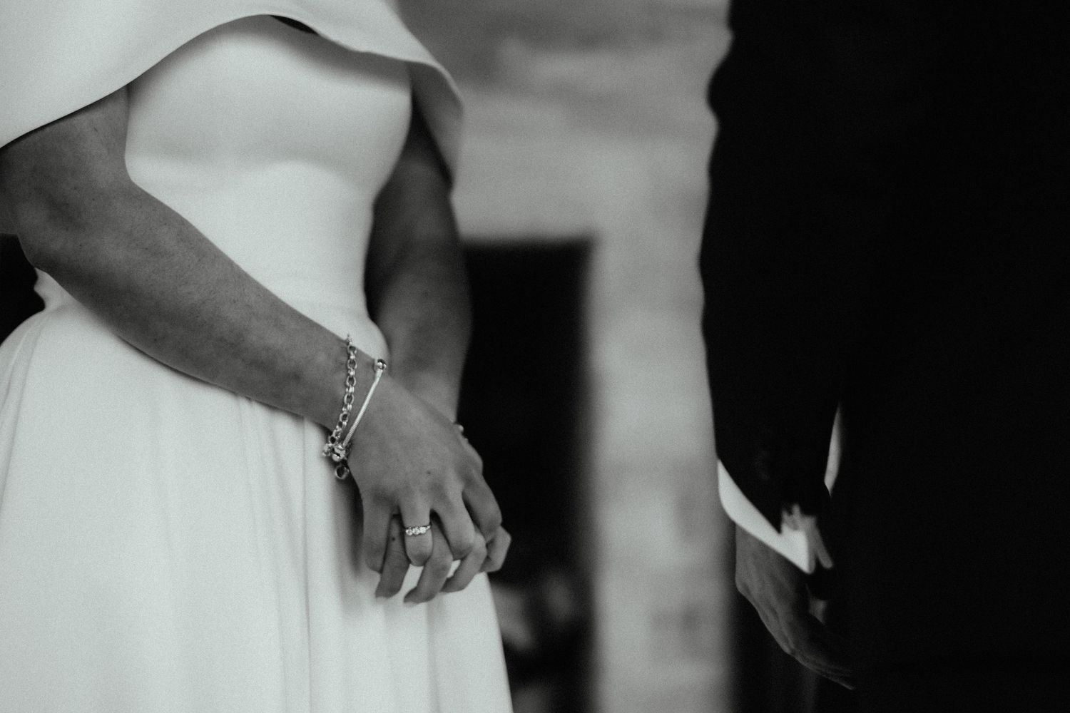Black and white close-up of a bride's hands wearing a bracelet and ring beside the groom during the ceremony.
