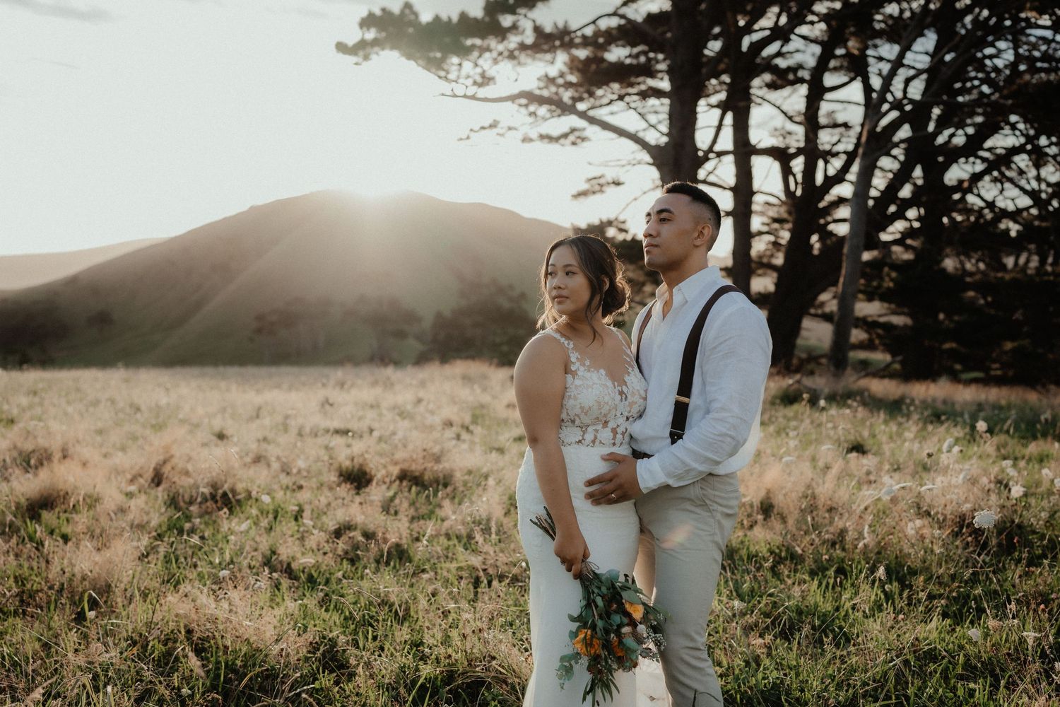 A bride and groom embrace in a golden meadow at sunset, with a bouquet in hand and hills glowing behind them.