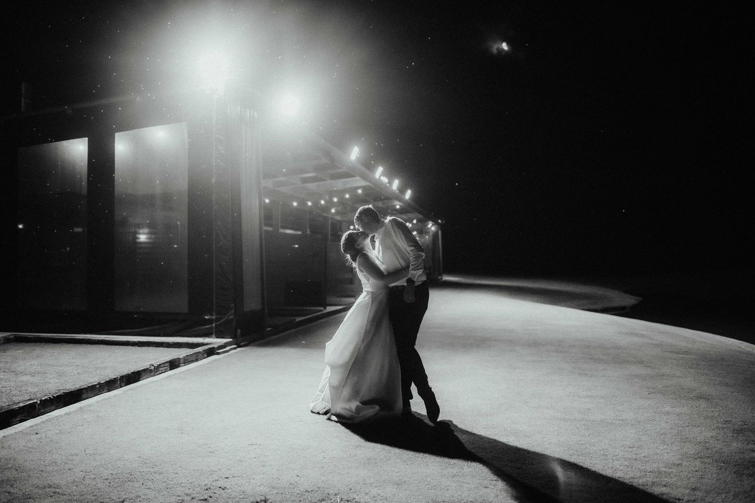 Bride and groom share a kiss on a dance floor at night under glowing lights in a dramatic black and white wedding photo.