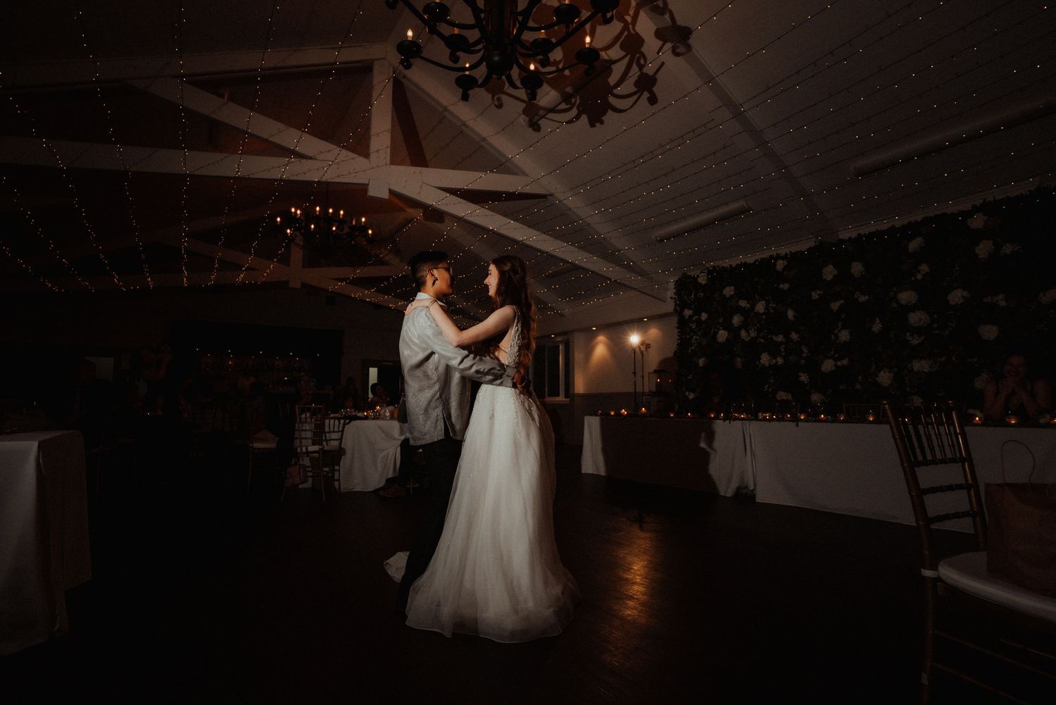Newlyweds share a romantic slow dance in a dimly lit reception hall with a chandelier and rustic wooden beams overhead.