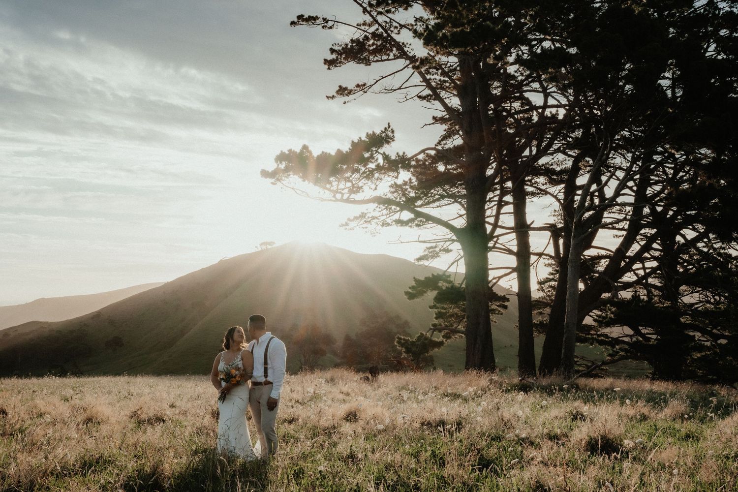 Bride and groom embrace in a grassy field beneath tall pine trees with sunburst over rolling hills.
