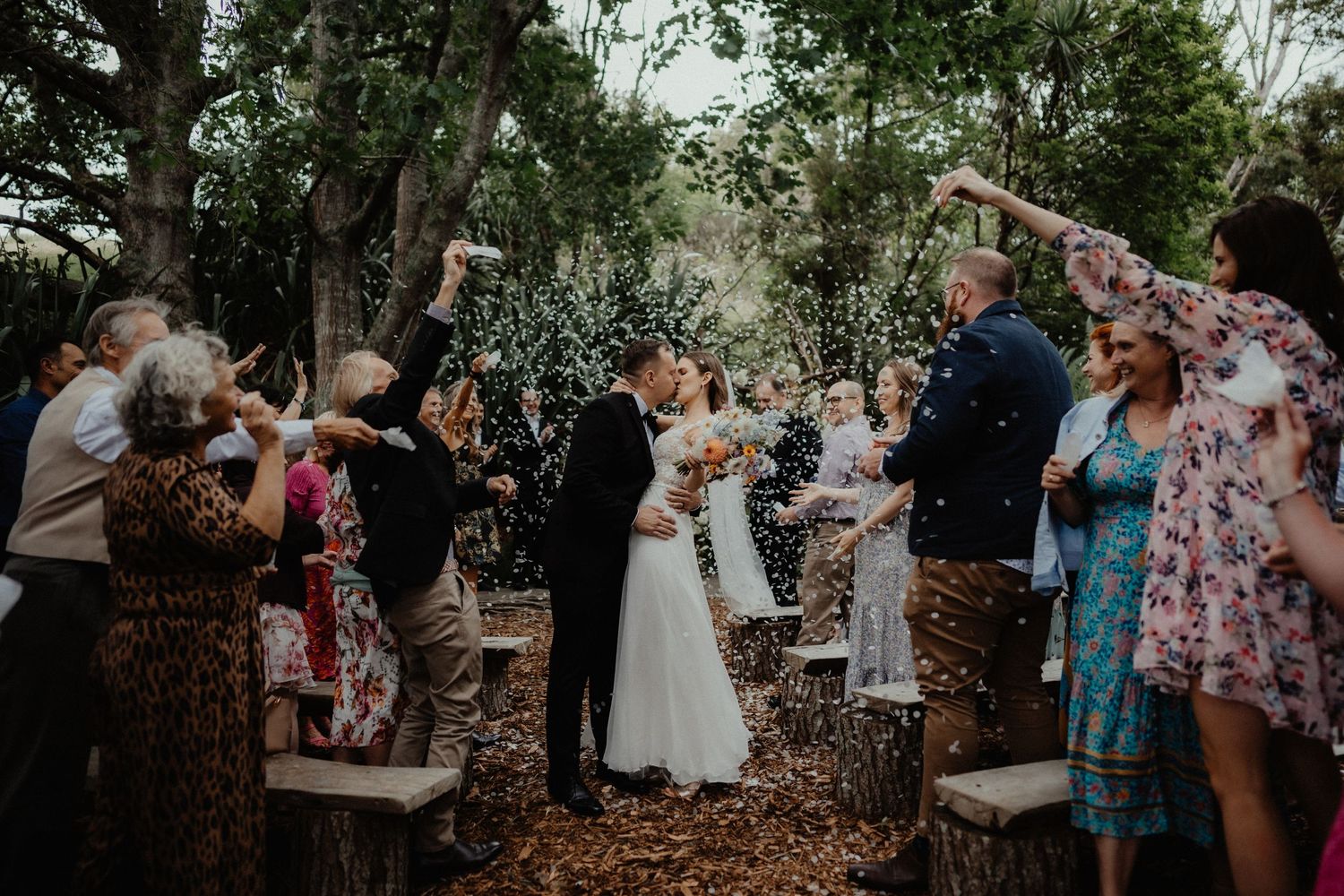 Guests cheer and toss confetti as the newlywed couple walk down the aisle after their outdoor woodland wedding ceremony.