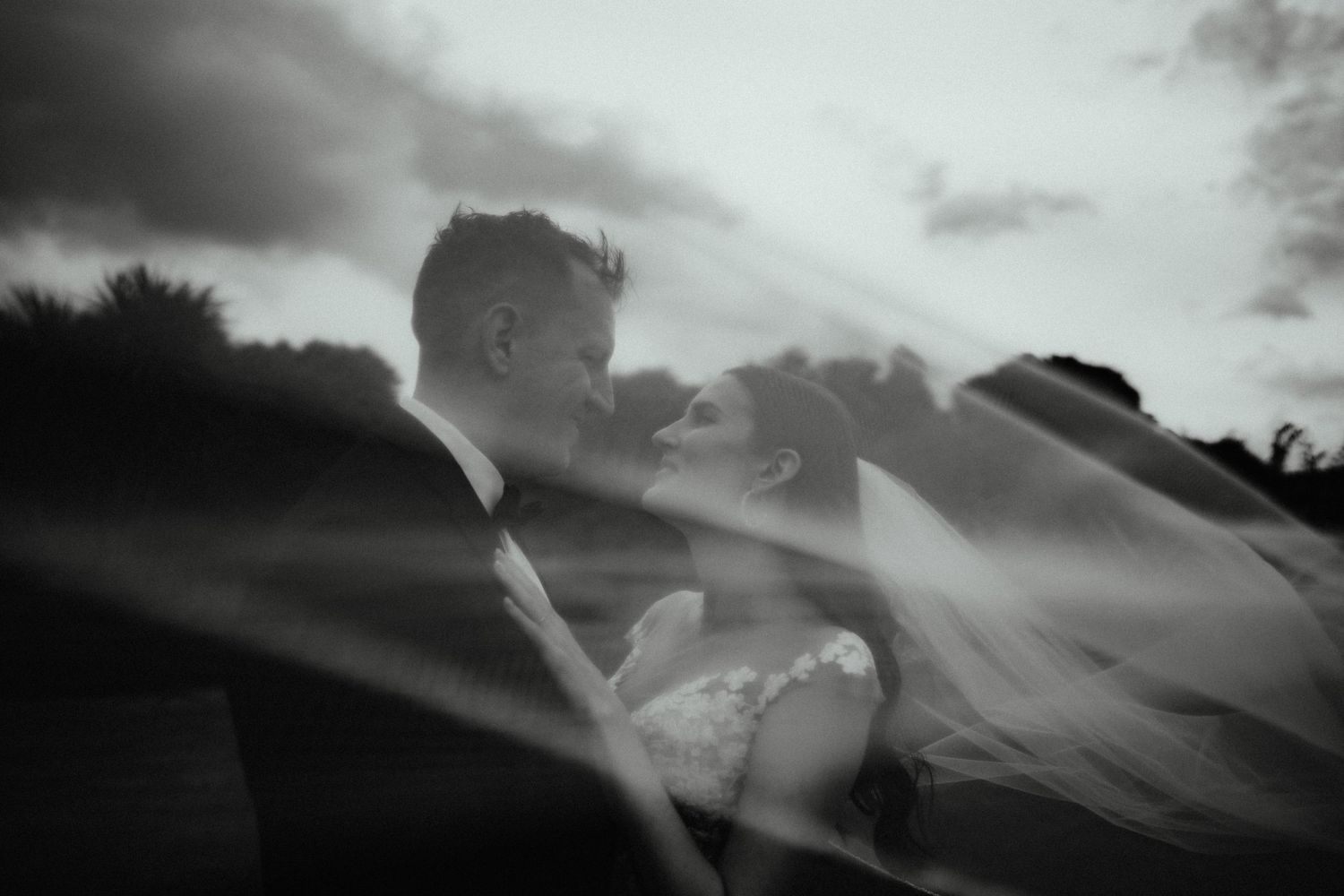 Black and white wedding photo of a couple nose to nose as a flowing veil sweeps across the dramatic cloudy sky.