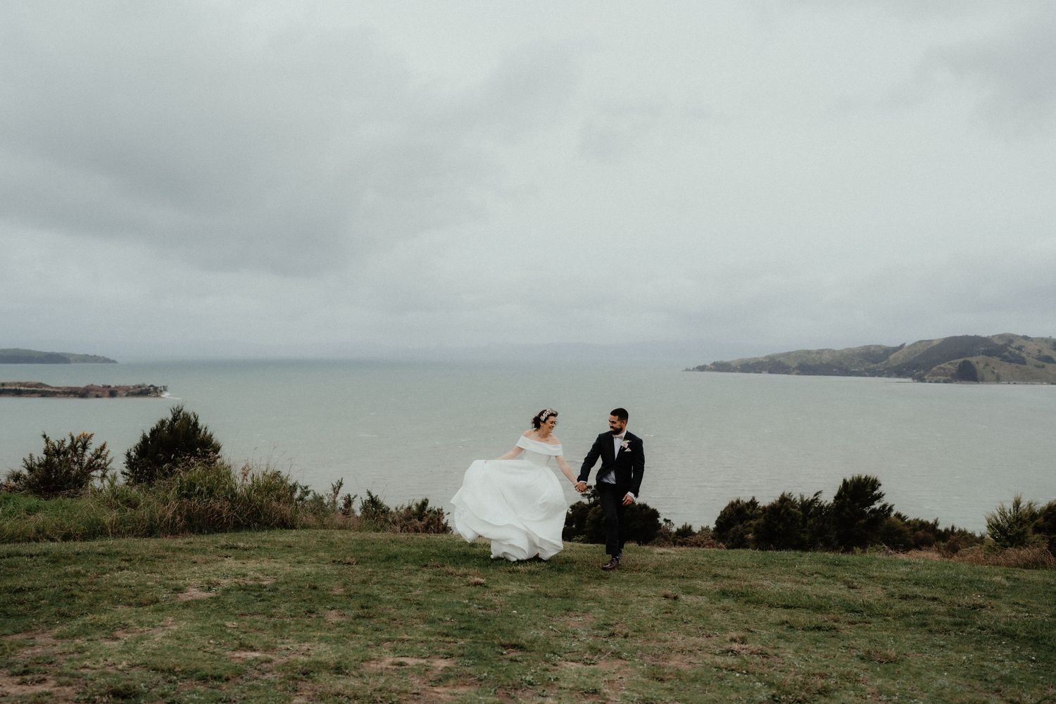 Bride in a flowing white gown and groom in a dark suit run hand in hand across a grassy hilltop overlooking the sea.