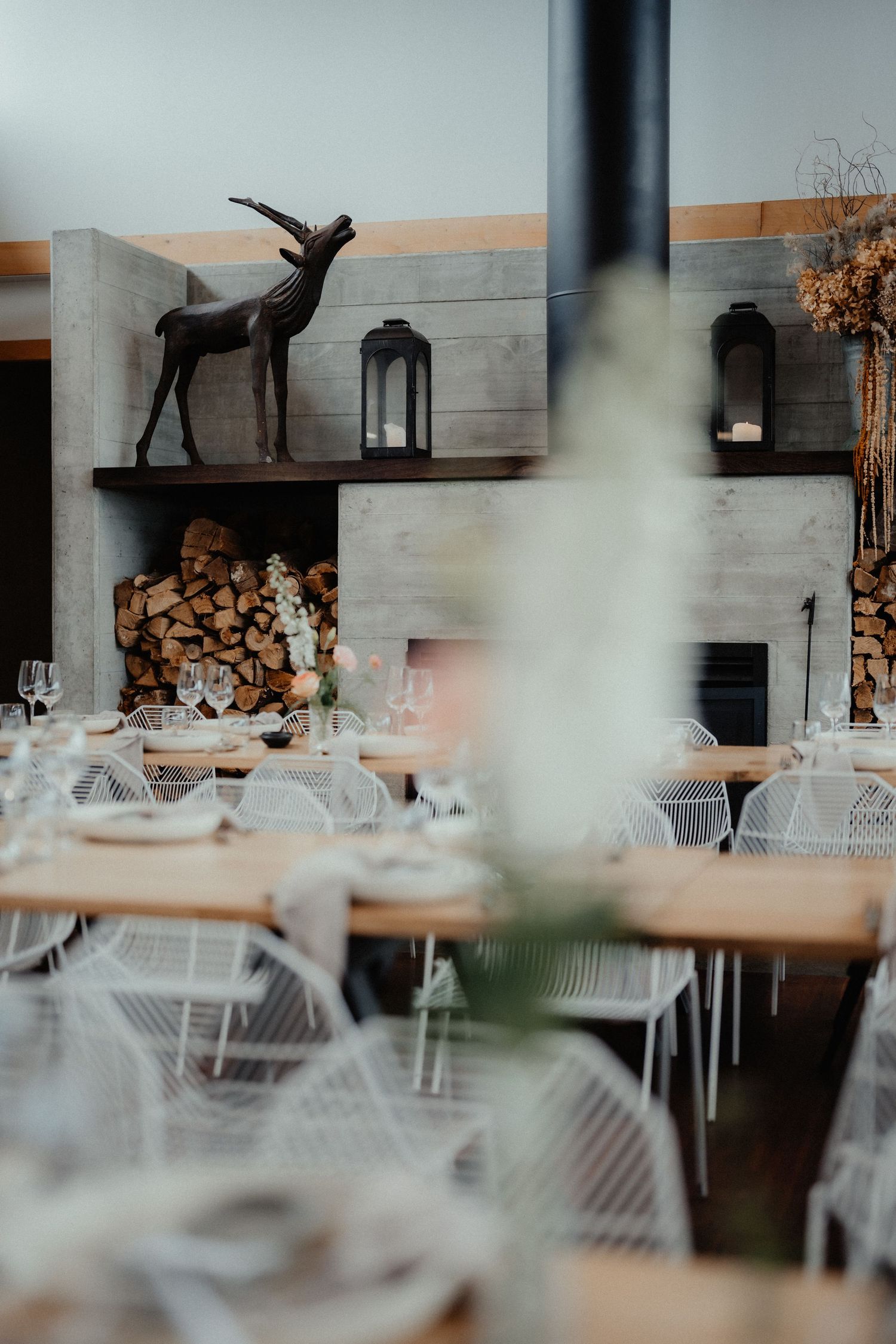 Rustic wedding reception tables with white wire chairs, a deer sculpture, lanterns and stacked firewood by a concrete fireplace.