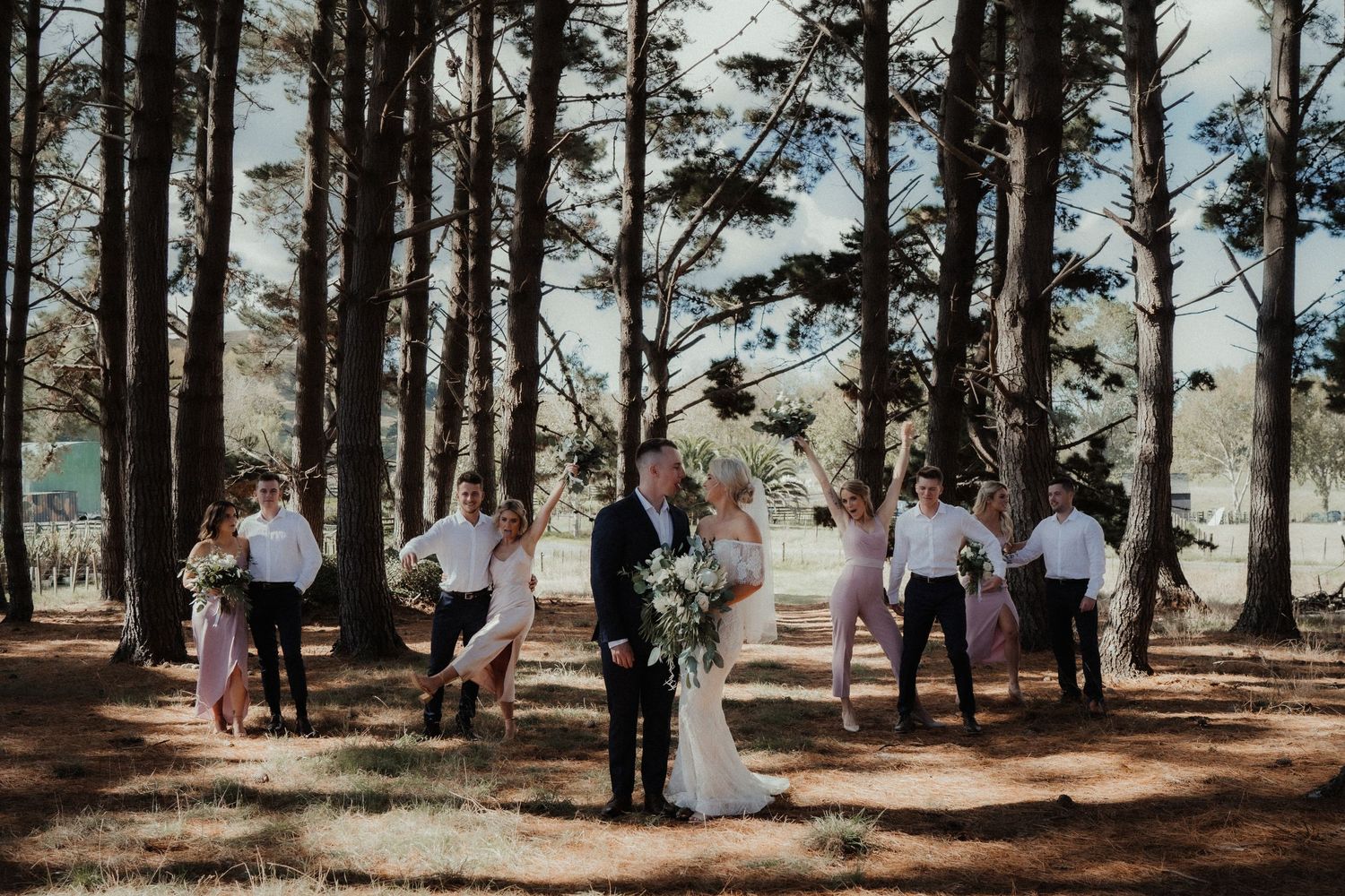 Bride and groom walk hand in hand with bridesmaids and groomsmen among tall pine trees at an outdoor forest wedding.