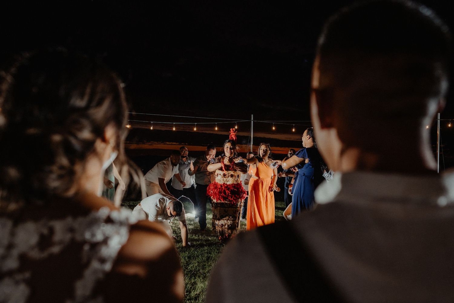 Wedding guests gather on a lawn at night to watch a lively performance under string lights.