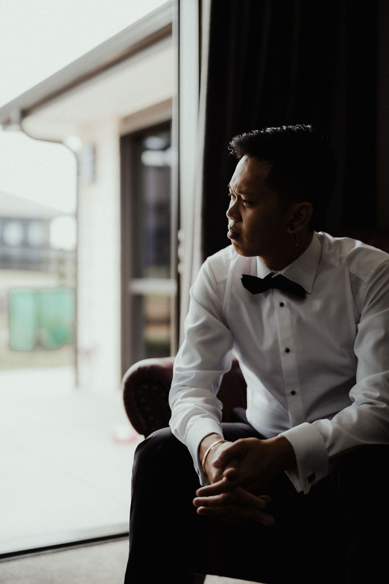 Groom in a white shirt and black bow tie sits thoughtfully beside a window, gazing outside before his wedding ceremony.