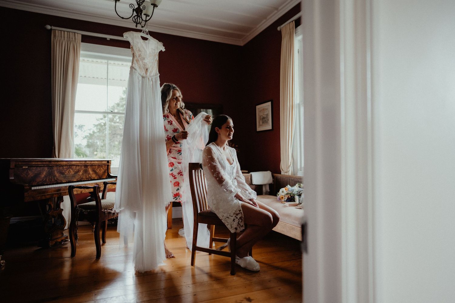 A bride sits by a window in a rustic room while a woman adjusts her hair and a white wedding dress hangs nearby.