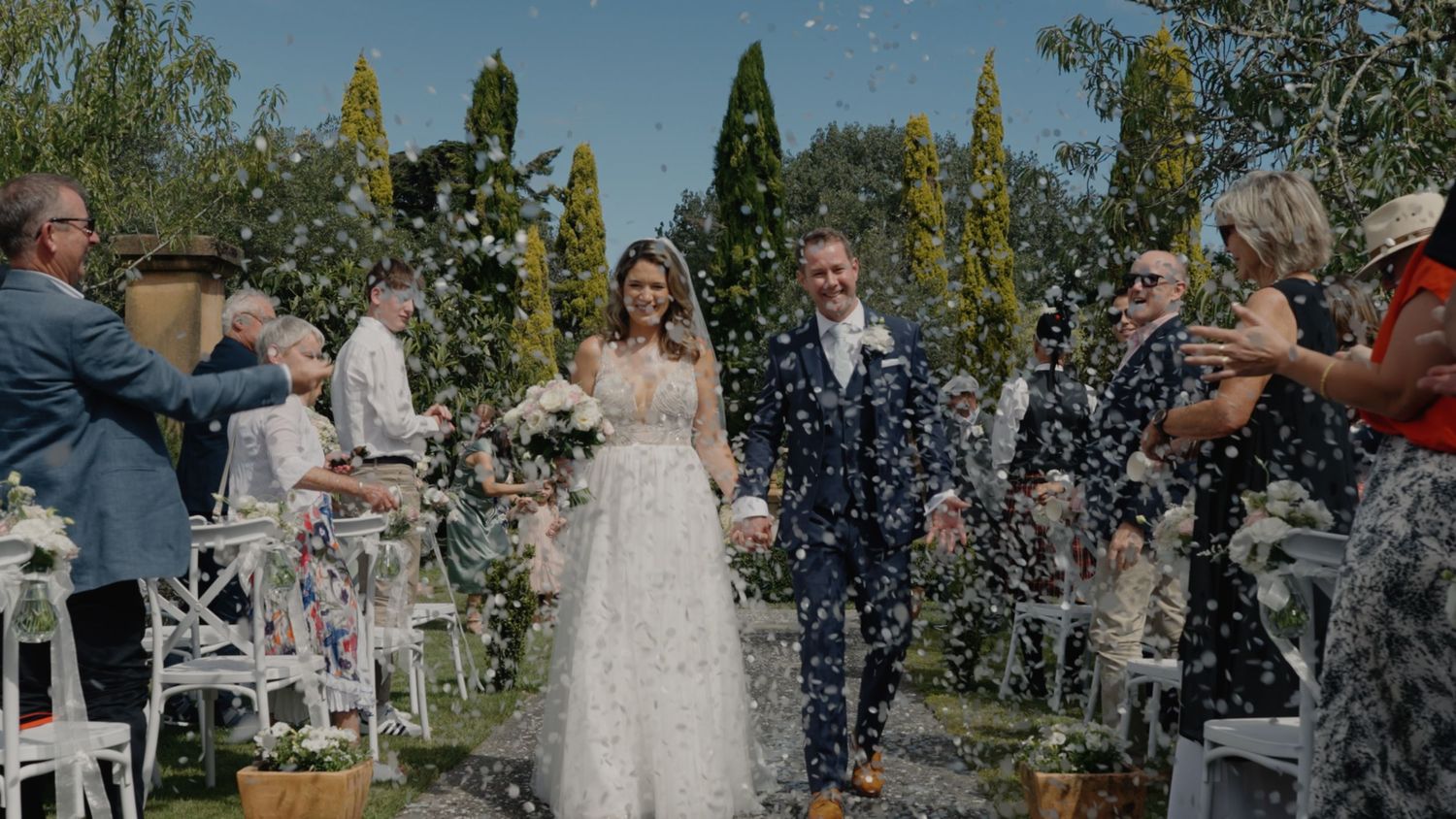Bride and groom walk hand in hand down the garden aisle as smiling wedding guests cheer and applaud around them.