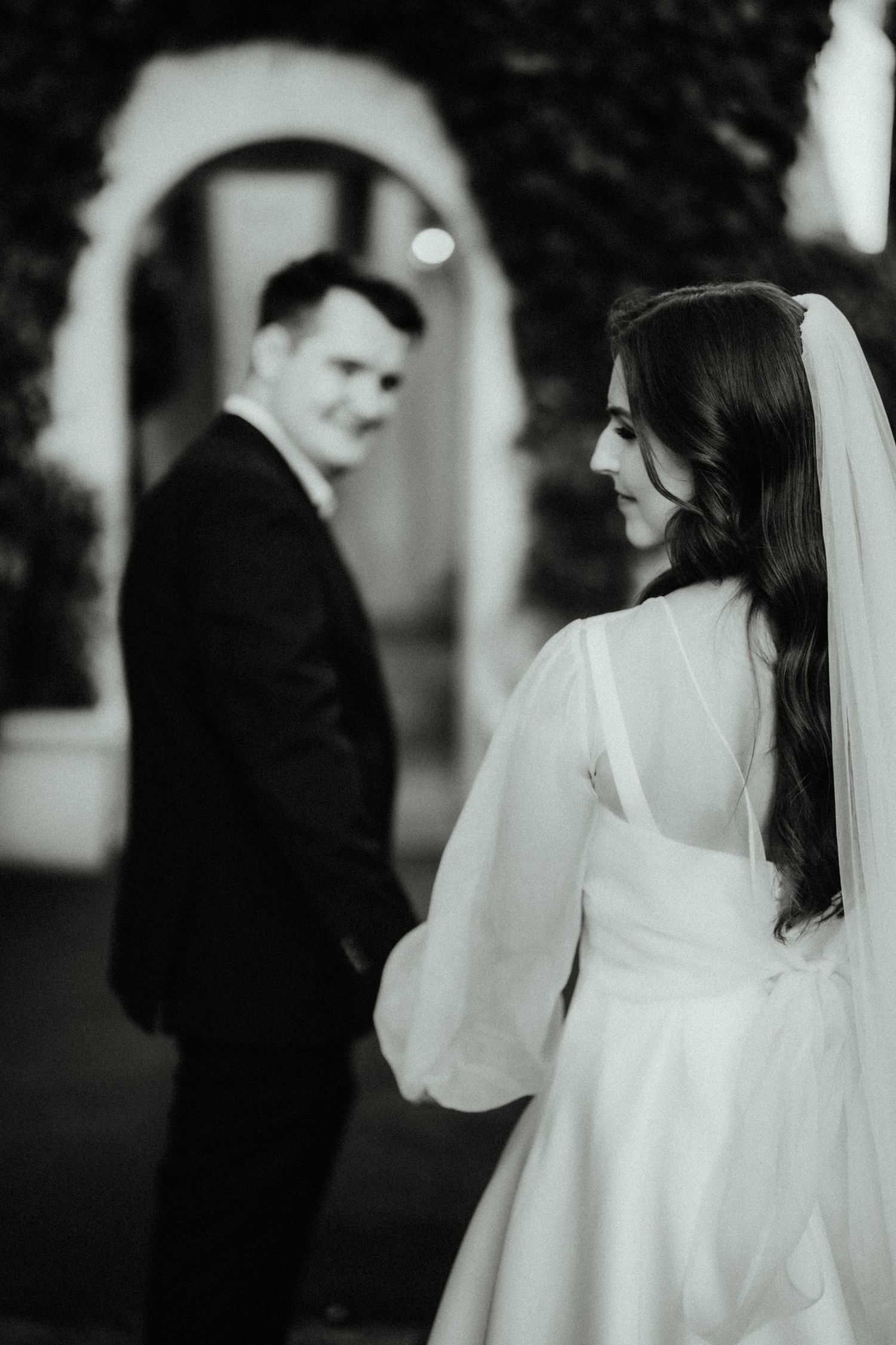Black and white wedding photo of a bride in a veil and sheer-sleeved gown glancing back at her smiling groom holding her hand.