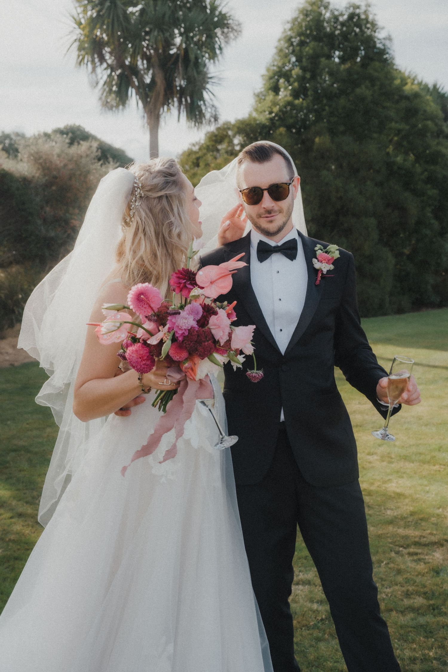 Bride in a white veil holding a pink bouquet stands beside a groom in sunglasses and tuxedo holding a champagne glass outdoors.