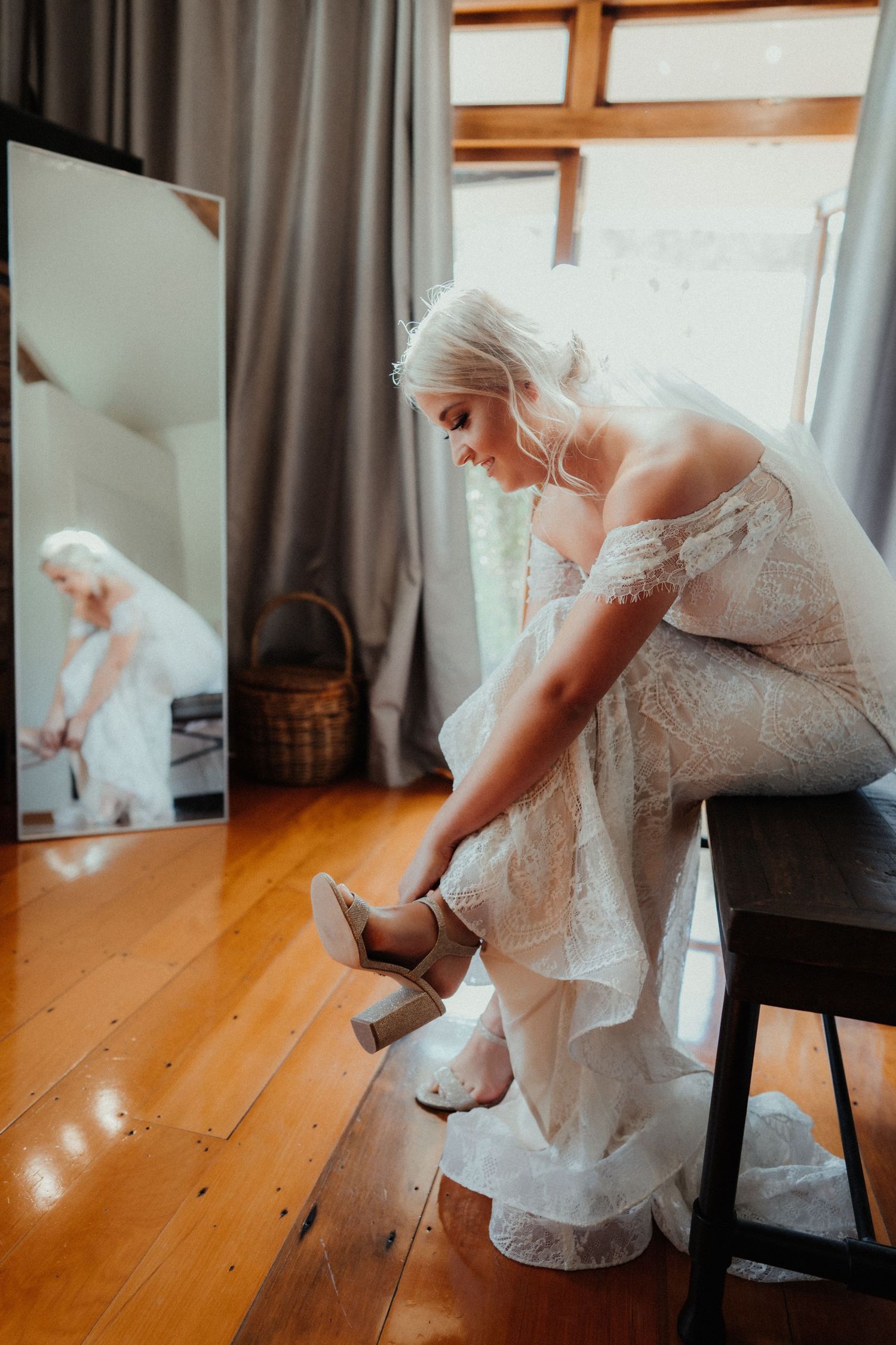 Bride in a lace off-the-shoulder wedding gown sits on a bench fastening her heeled sandal beside a mirror and window.