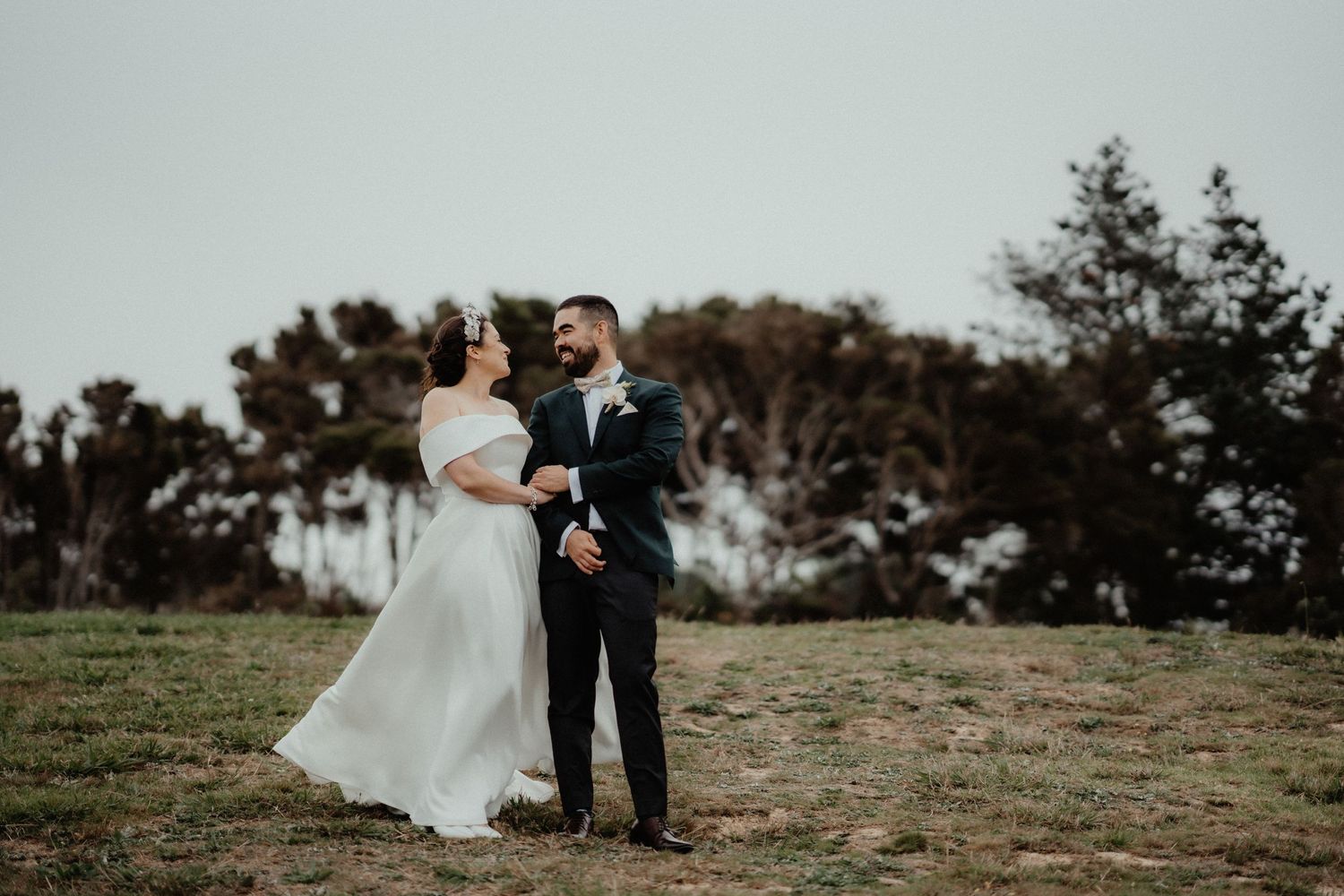Bride in a flowing white gown and groom in a dark suit embrace on a grassy hillside with trees under an overcast sky.