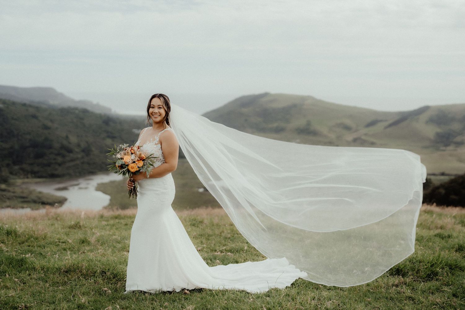 Smiling bride in a fitted white gown holds an orange bouquet as her long veil blows in the wind on a green hillside.