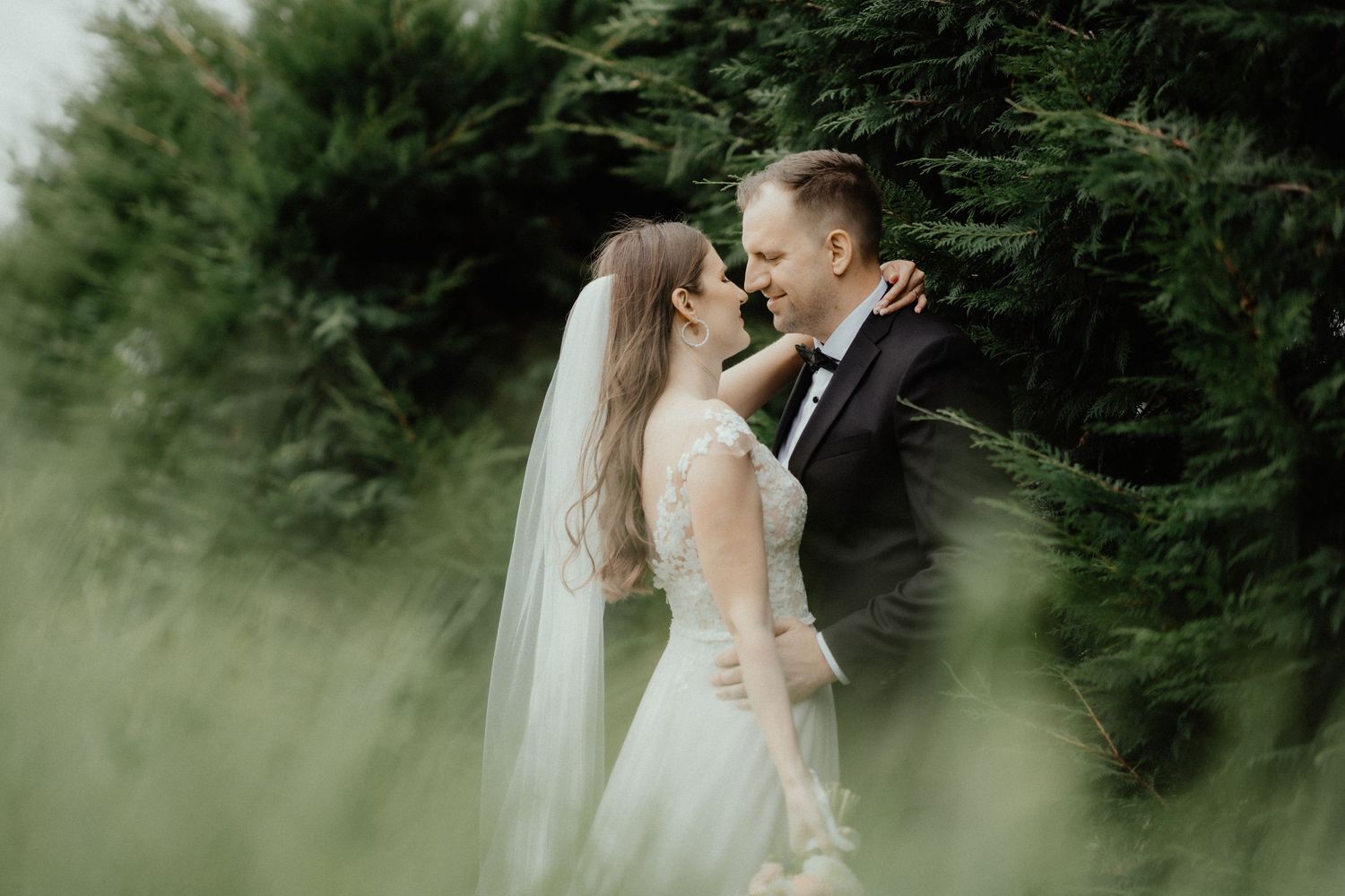 Bride and groom share a tender moment embracing beside tall green hedges, the bride's long veil trailing in the soft light.