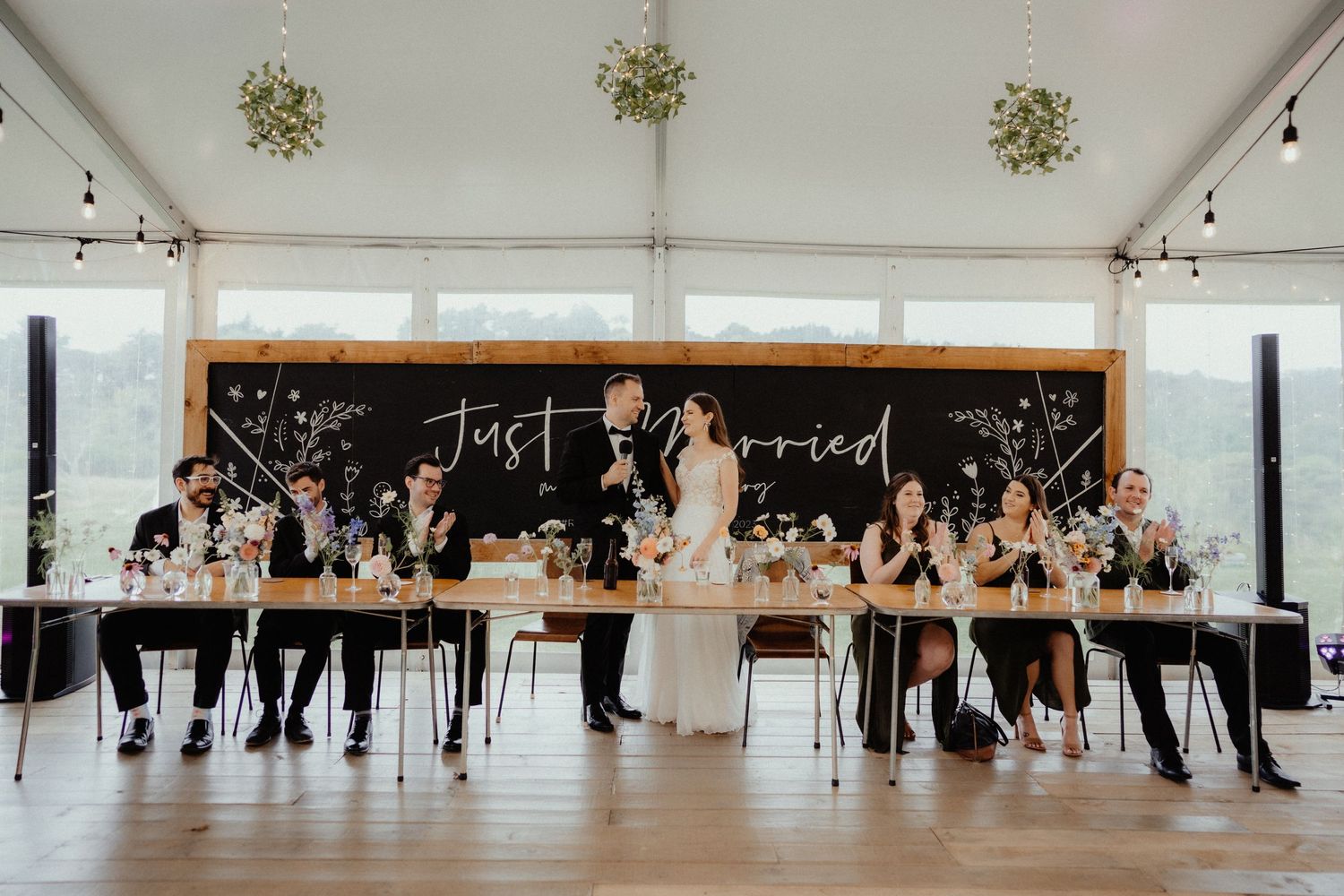 Bride and groom stand behind a long reception table with their wedding party beneath a Just Married chalkboard sign.