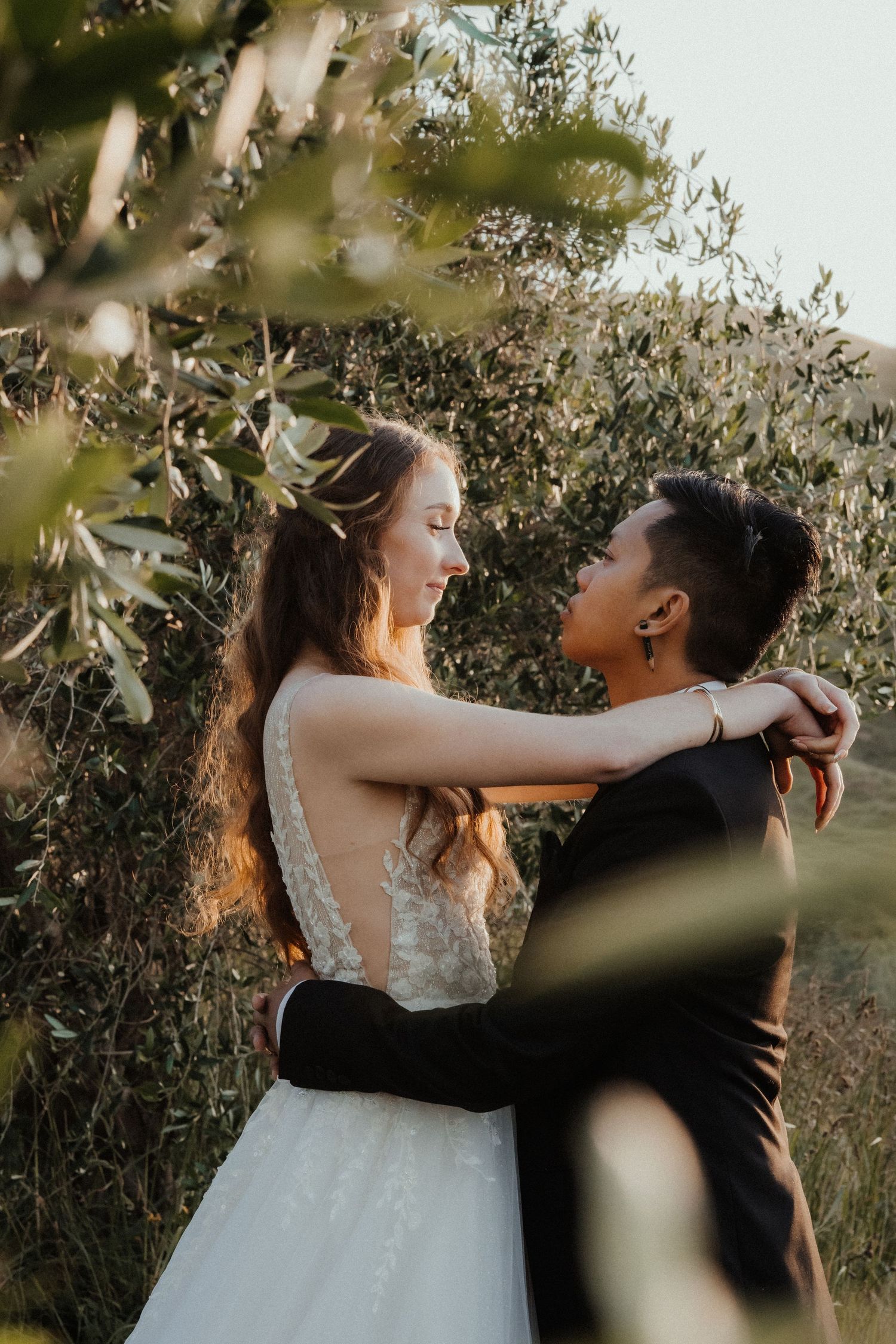 A bride in a lace-back wedding dress and groom in a dark suit embrace face to face amid olive branches in warm evening light.