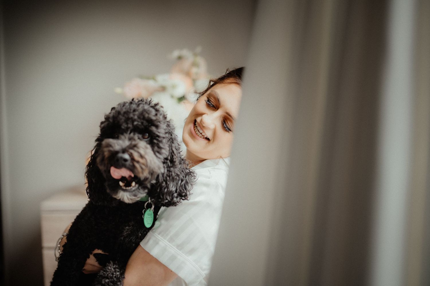 Smiling bride in a robe cuddles a fluffy black dog with a green tag in a softly lit room with flowers behind her.