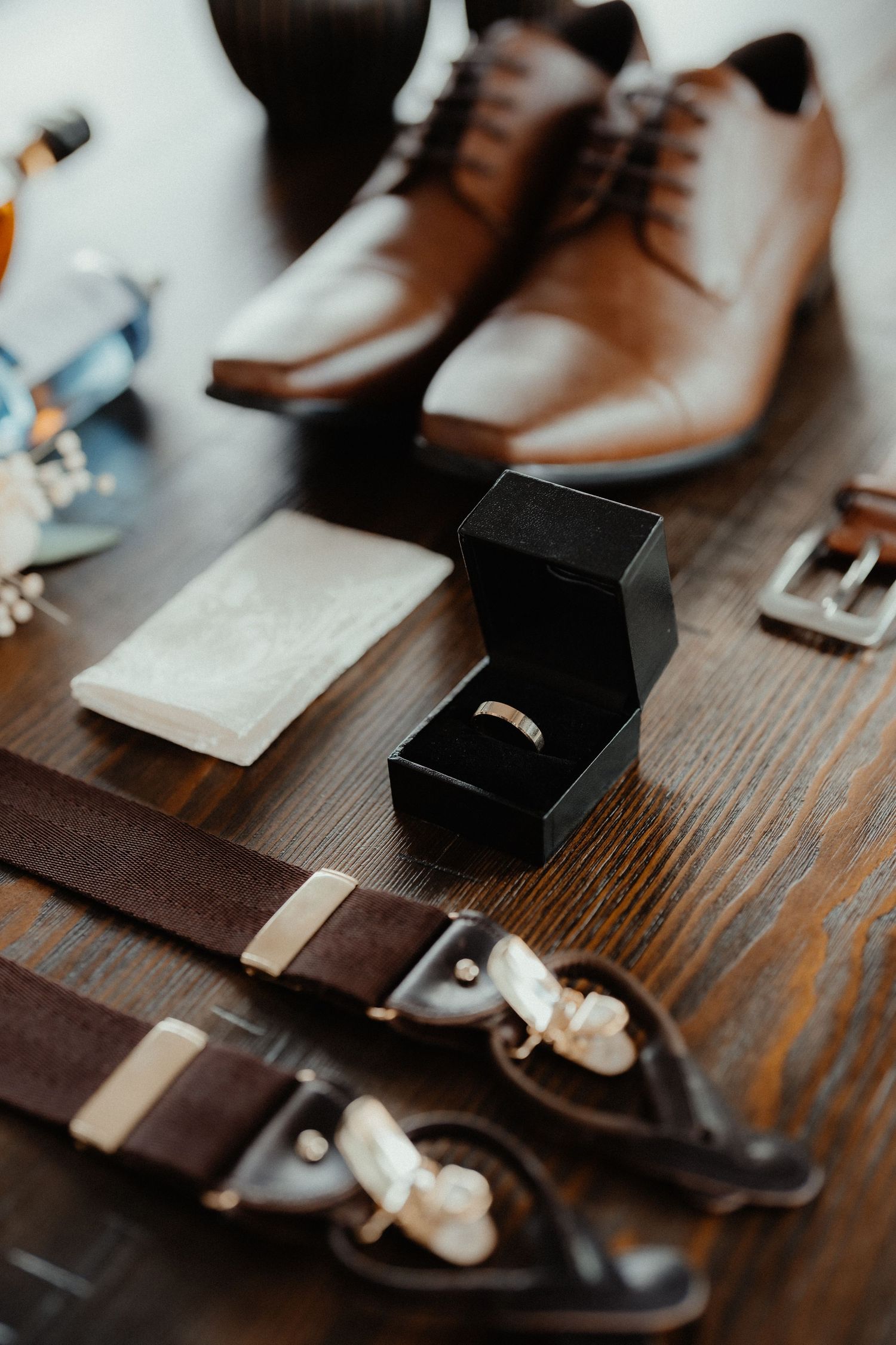 Wedding day flat lay with brown leather shoes, a ring box holding a gold band, suspenders, a belt buckle and a pocket square.