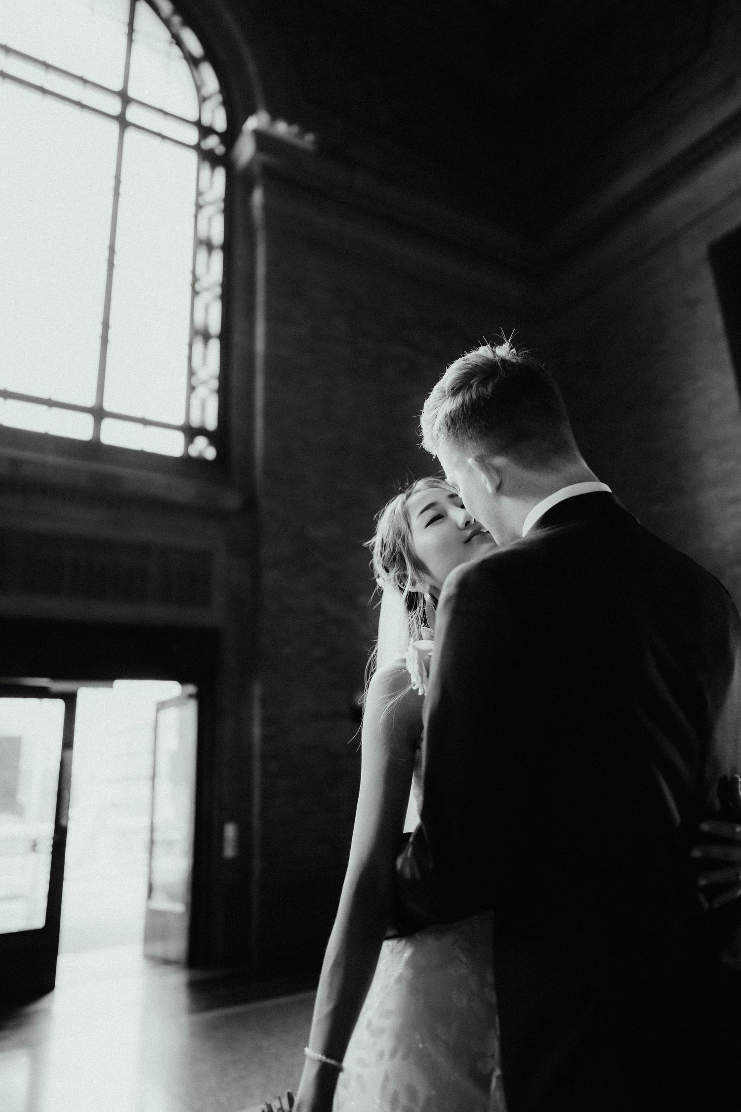 Bride and groom share a kiss in a grand hall with tall arched windows in a black and white wedding photo.
