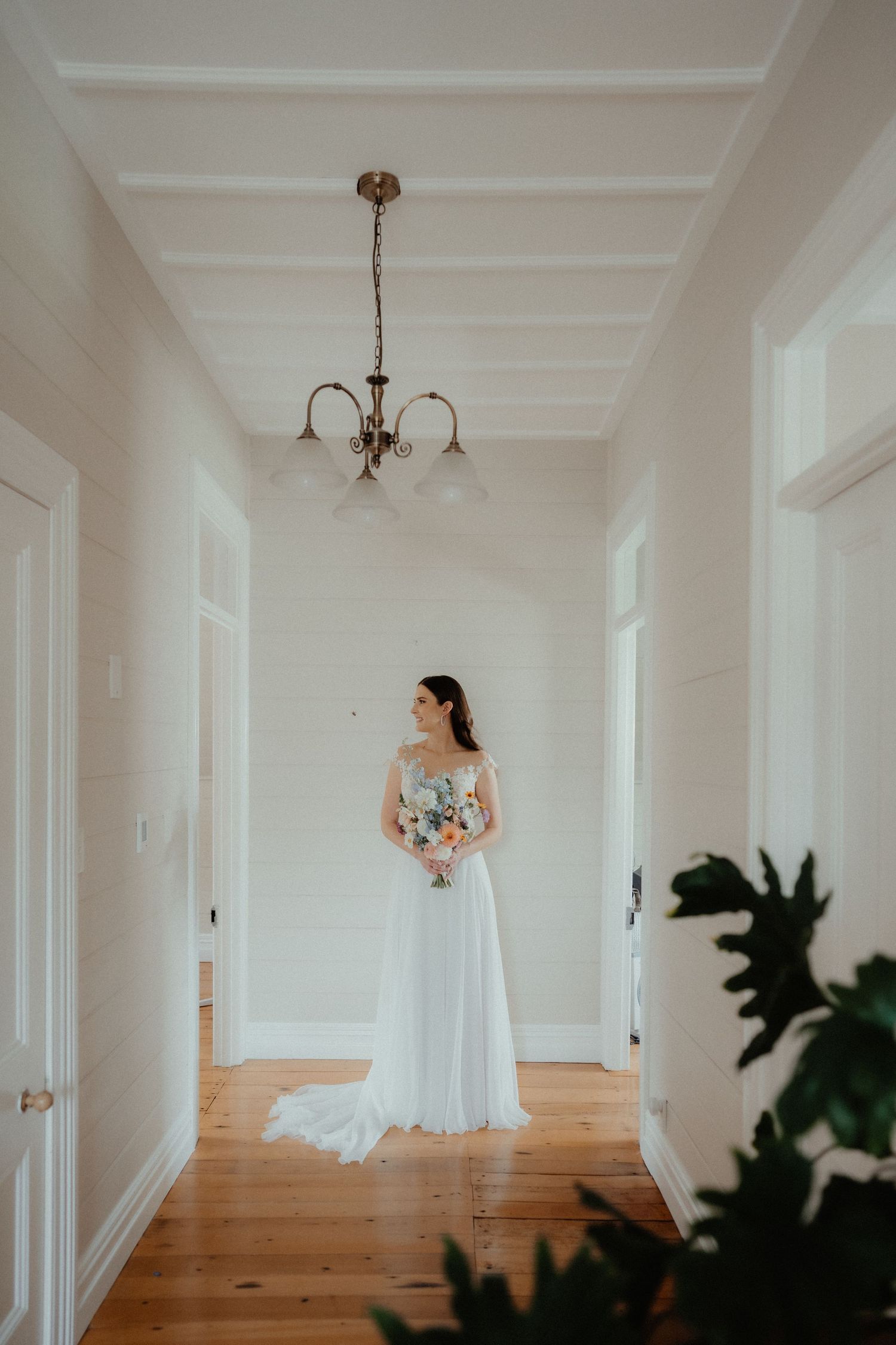 Bride in a flowing white gown holds a bouquet while standing in a bright hallway with wooden floors and a chandelier.