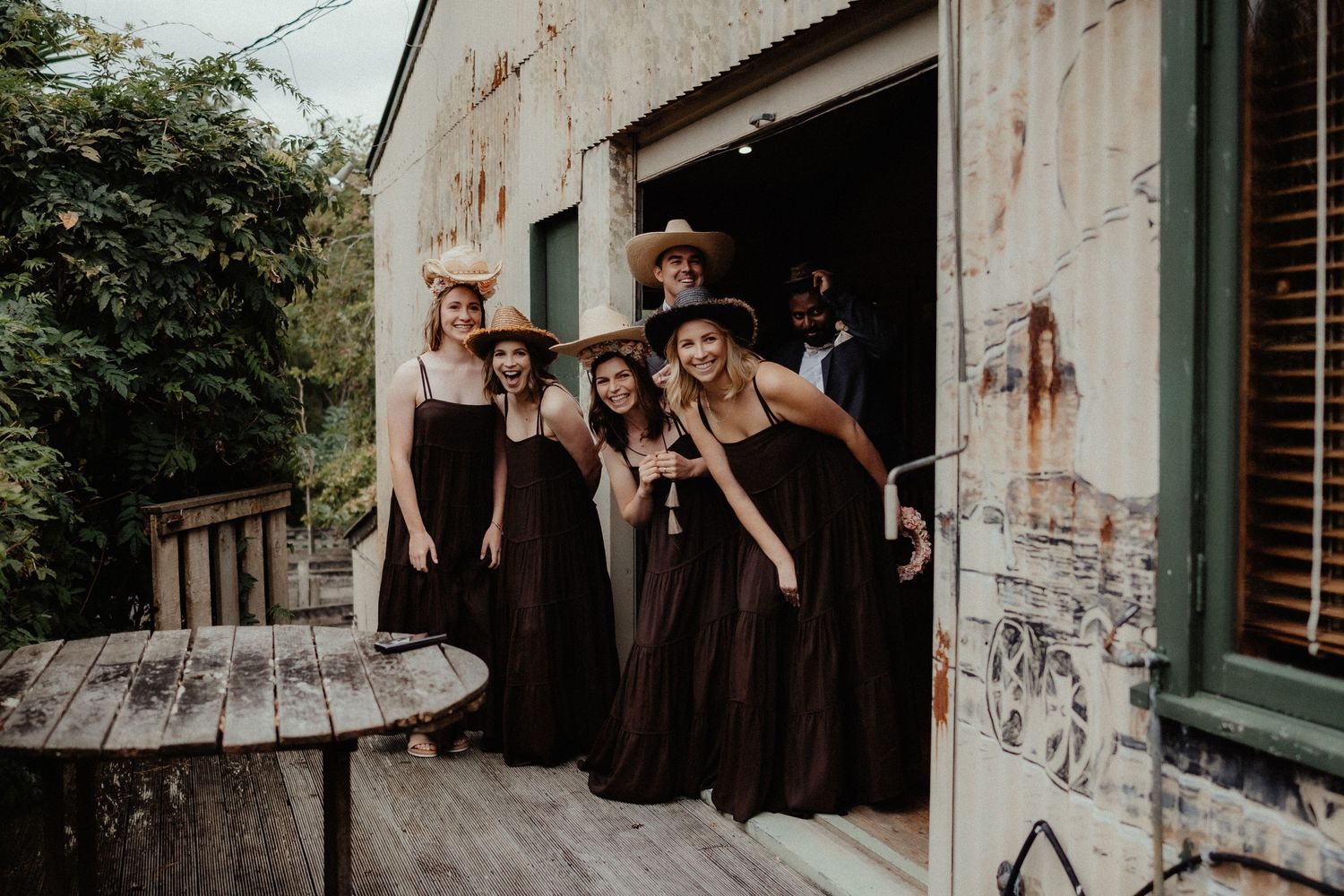Bridesmaids in long black dresses and wide-brimmed hats pose together on a rustic wooden porch outside a weathered building.